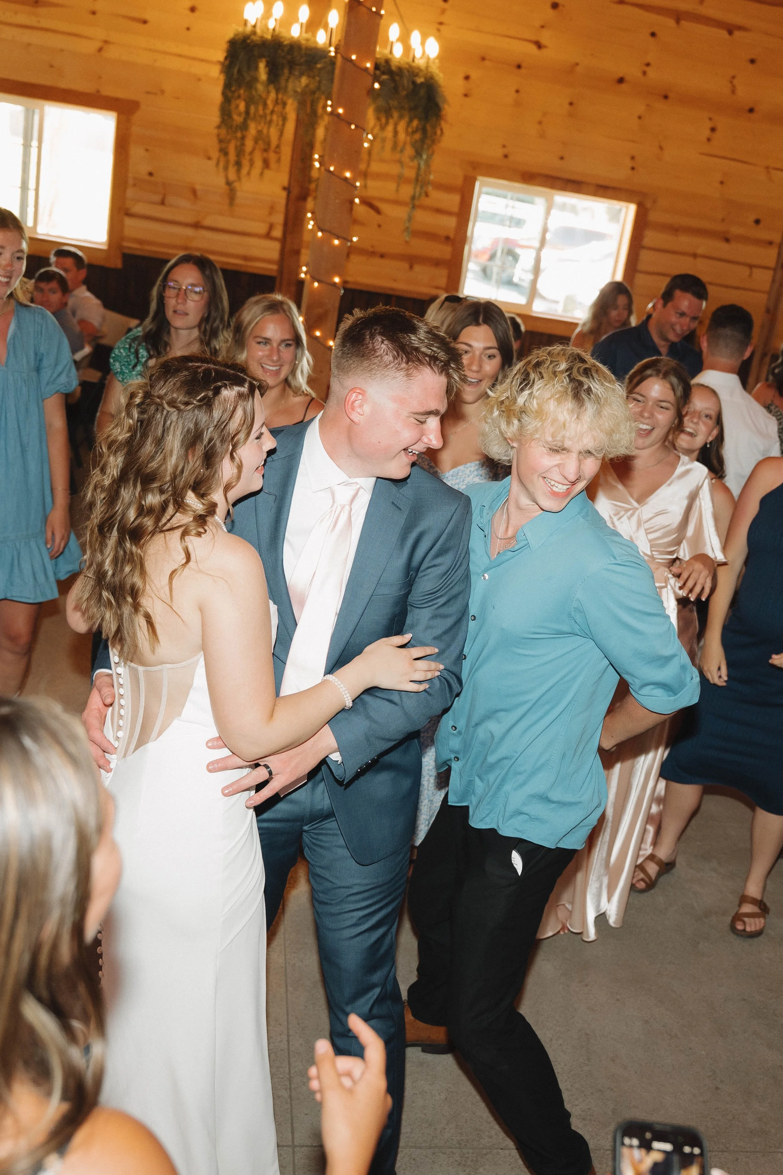 A group of people dancing and celebrating at a wedding reception inside a wooden hall with string lights and hanging greenery.