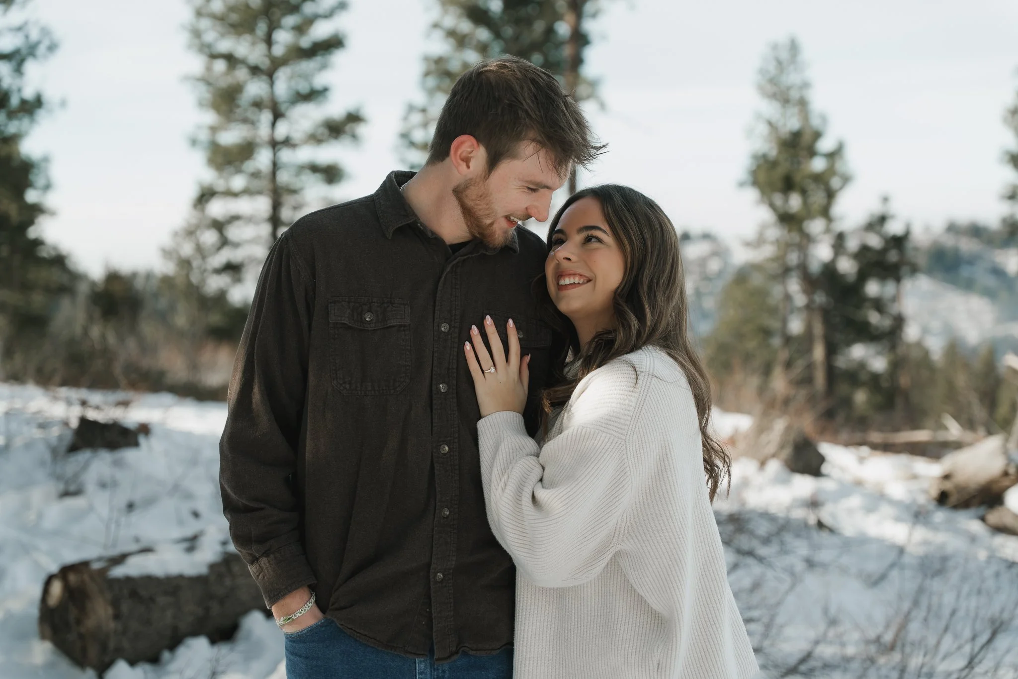 A couple standing close together outdoors in a snowy forest, smiling at each other, with the woman touching the man's chest and wearing an engagement ring.