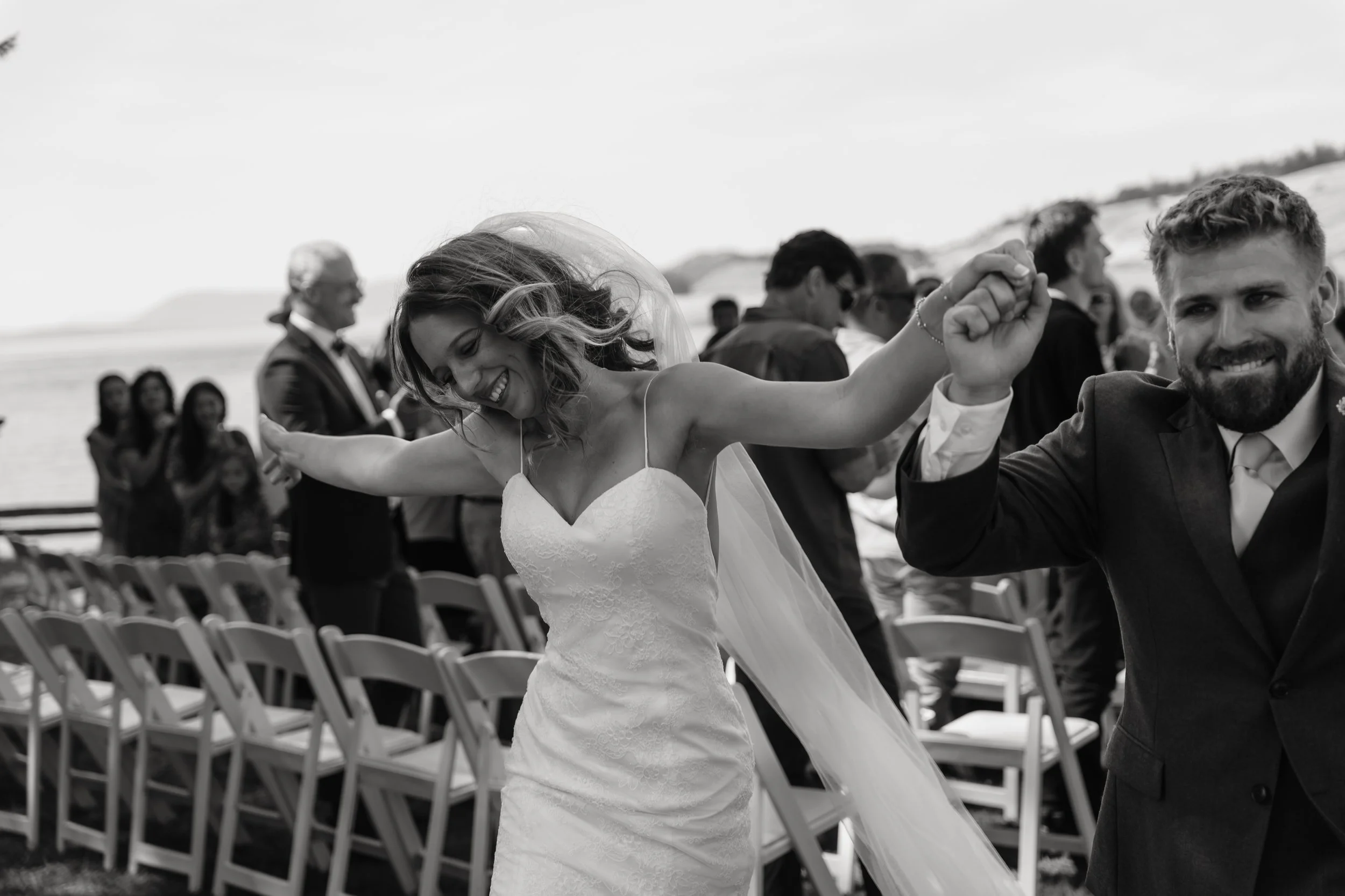 A black-and-white photo of a bride and groom dancing and smiling at their outdoor wedding, with guests in the background near chairs and a seaside landscape.