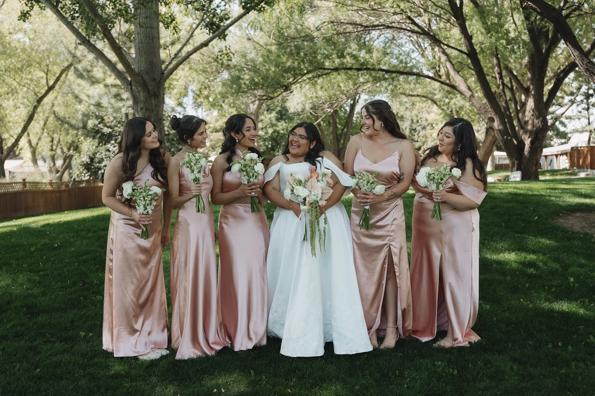 A bride in a white wedding gown standing in the center, surrounded by six bridesmaids dressed in matching blush pink satin dresses, all holding small bouquets of white flowers, outdoors with green grass and large trees in the background.