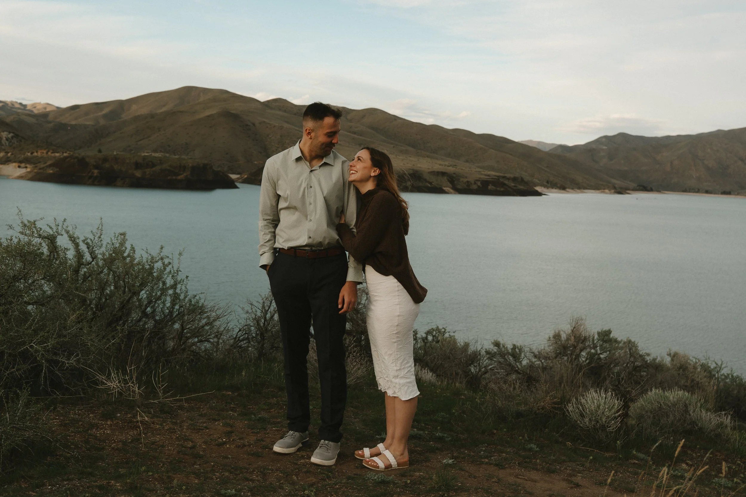 A couple standing on a hillside near a lake, smiling and looking at each other, with hills in the background.
