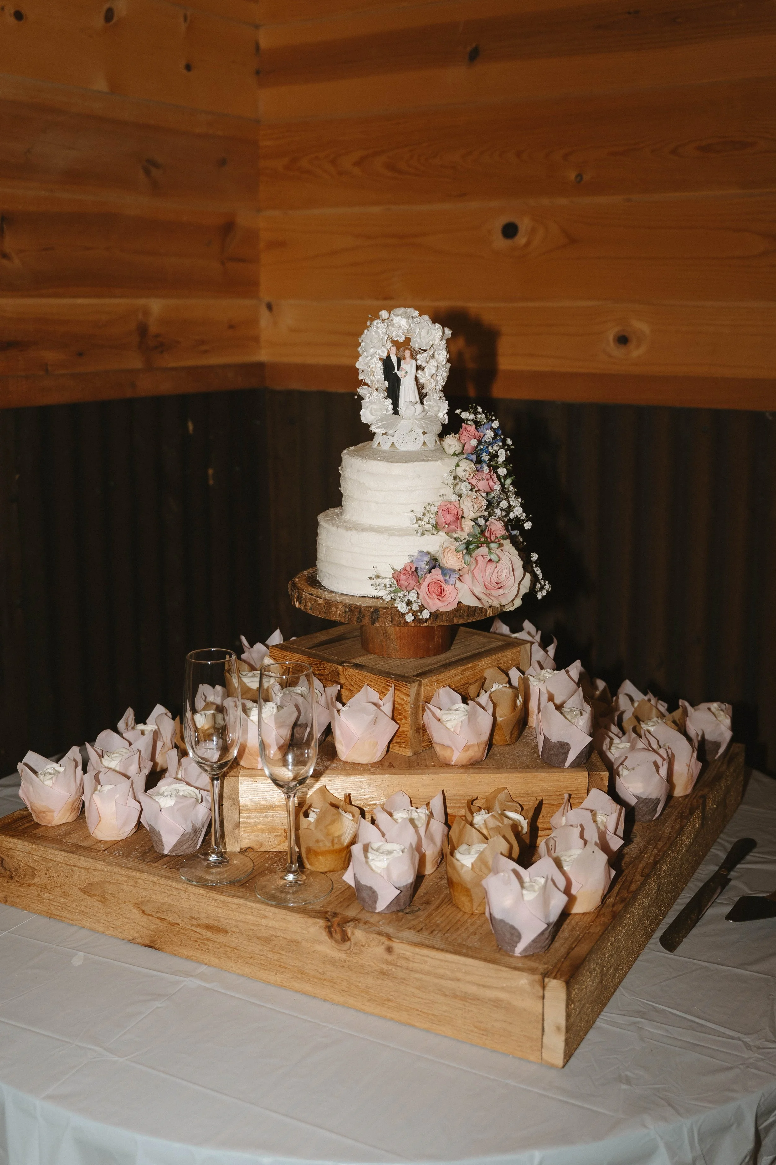 A wedding cake decorated with flowers and a miniature bride figurine on top, surrounded by small cupcake-like desserts in colored wrappers, and two empty wine glasses on a wooden tray.