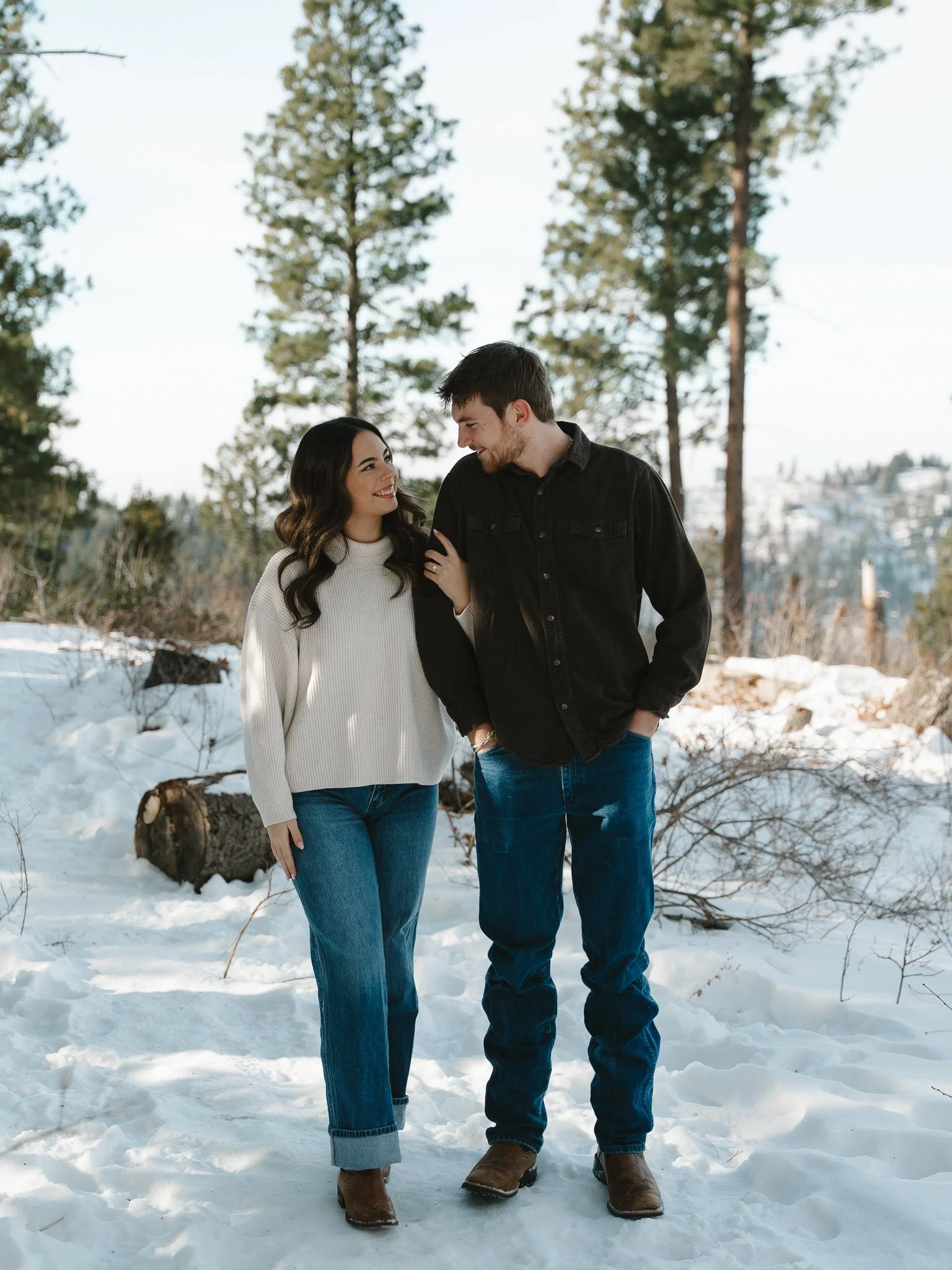 A young couple standing close together on snowy ground in a forest, smiling and looking at each other.