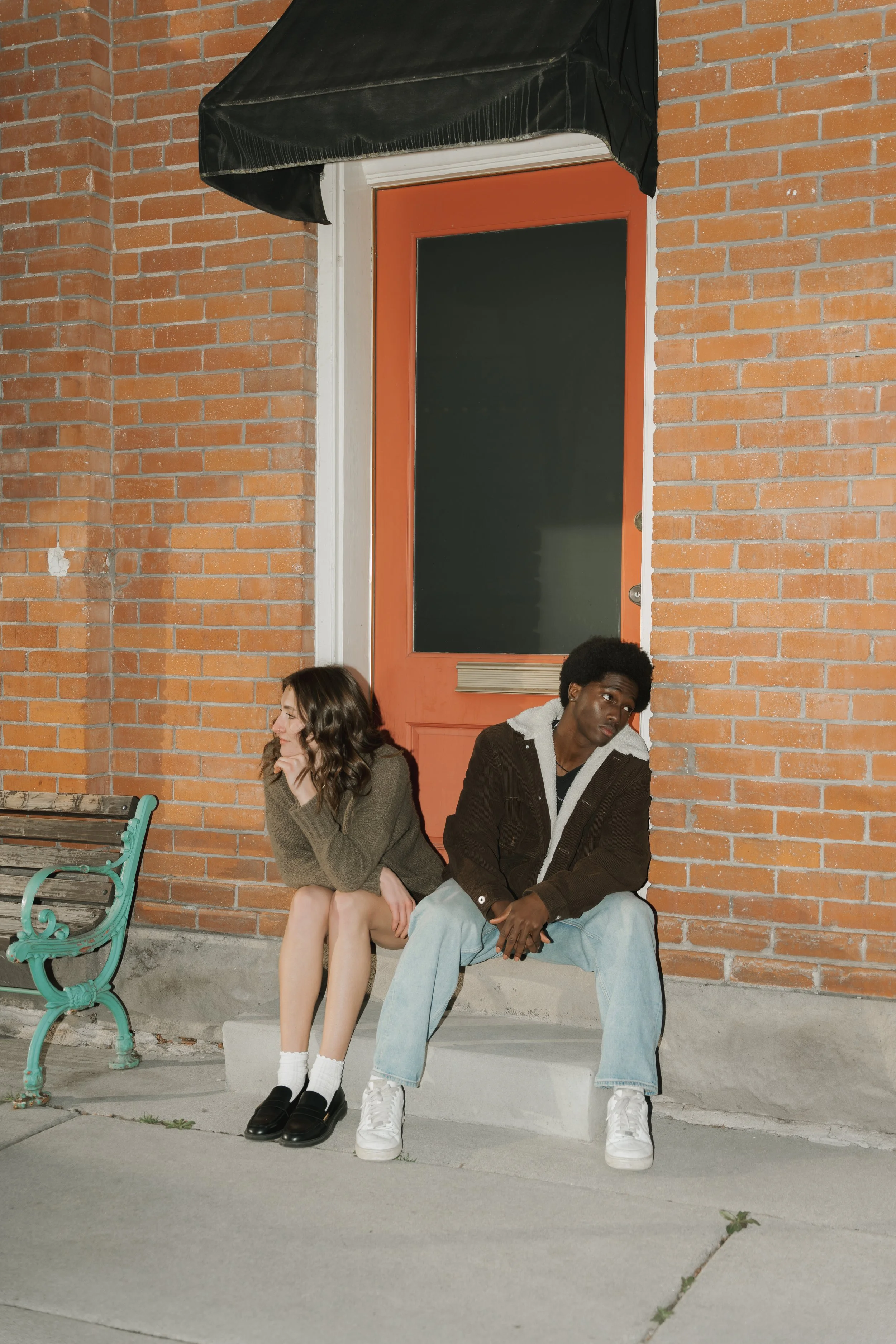 A young woman and a young man sit on the sidewalk against a brick wall with a red door behind them. The woman is smiling and looking to her left, while the man looks downward with a thoughtful expression.