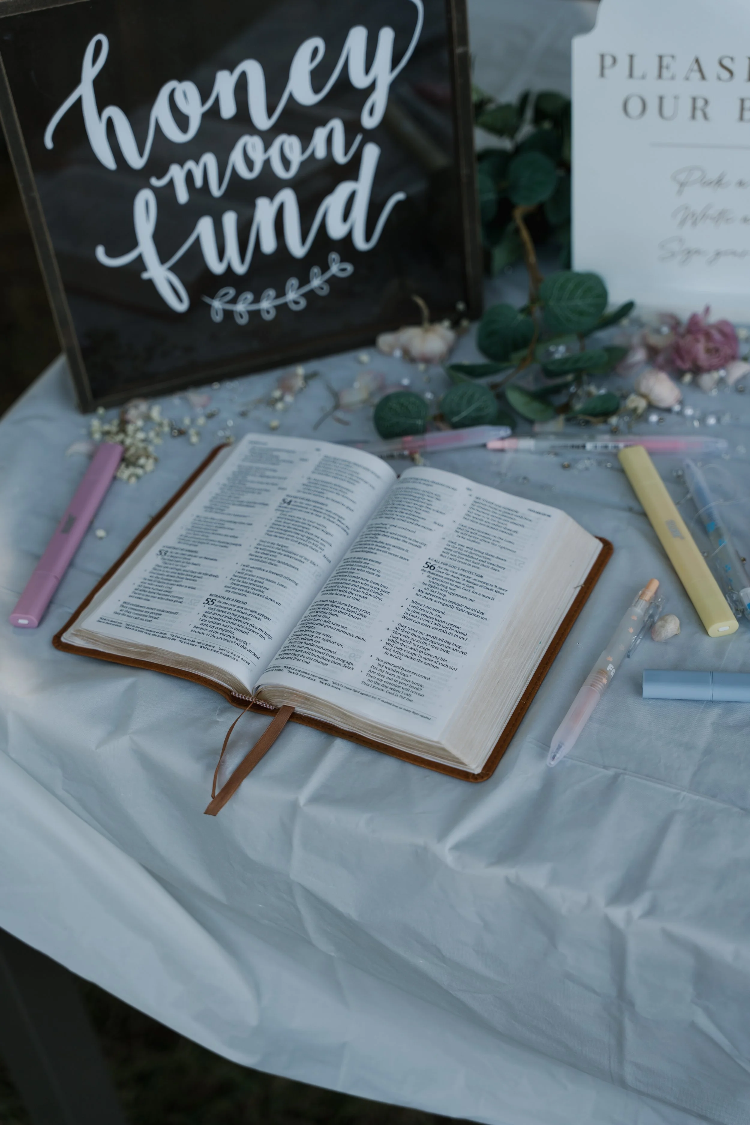 Open Bible on a table with various pens around, floral decorations, and a sign that says 'honey moon fund'.