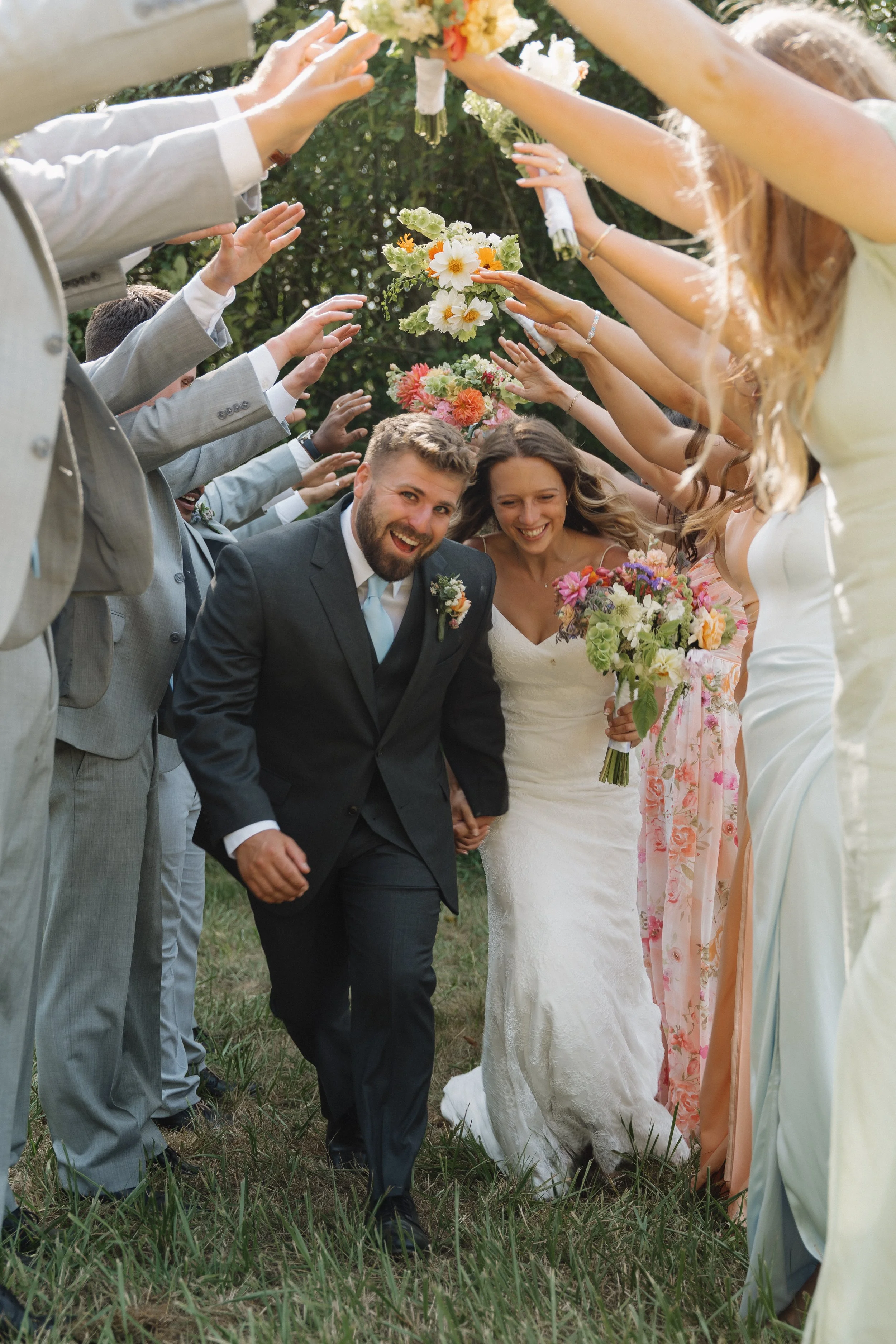 A newlywed couple walking through a tunnel of friends and family holding flowers at an outdoor wedding.