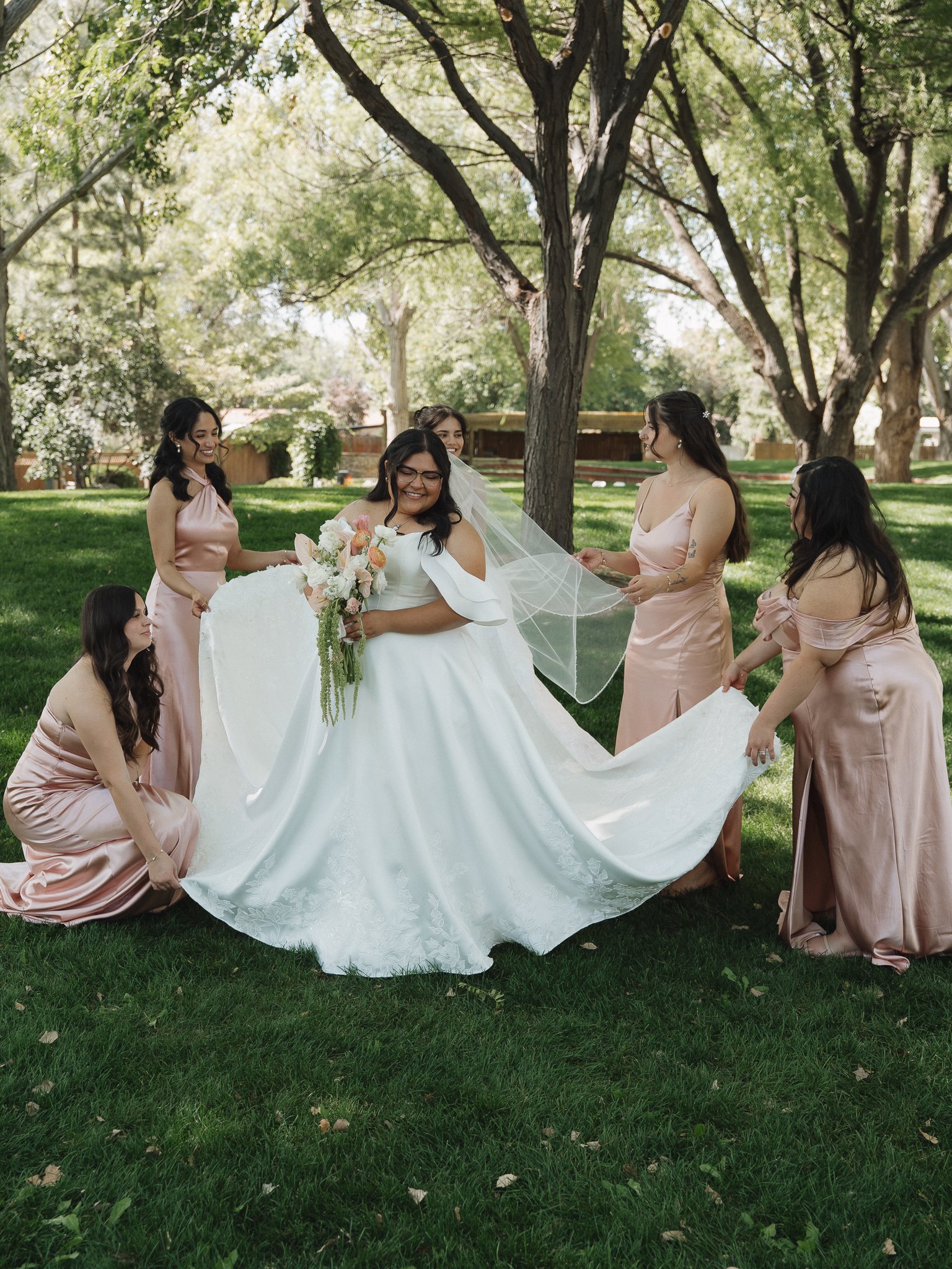 A bride in a white wedding dress holding a bouquet of flowers is surrounded by bridesmaids in pink dresses, outdoors under large trees on a grassy lawn.