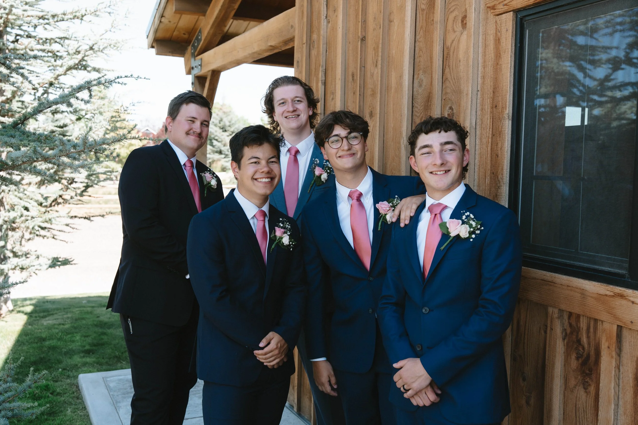 Six young men dressed in formal suits with pink ties and boutonnieres, standing outside near a wooden building, smiling at the camera.