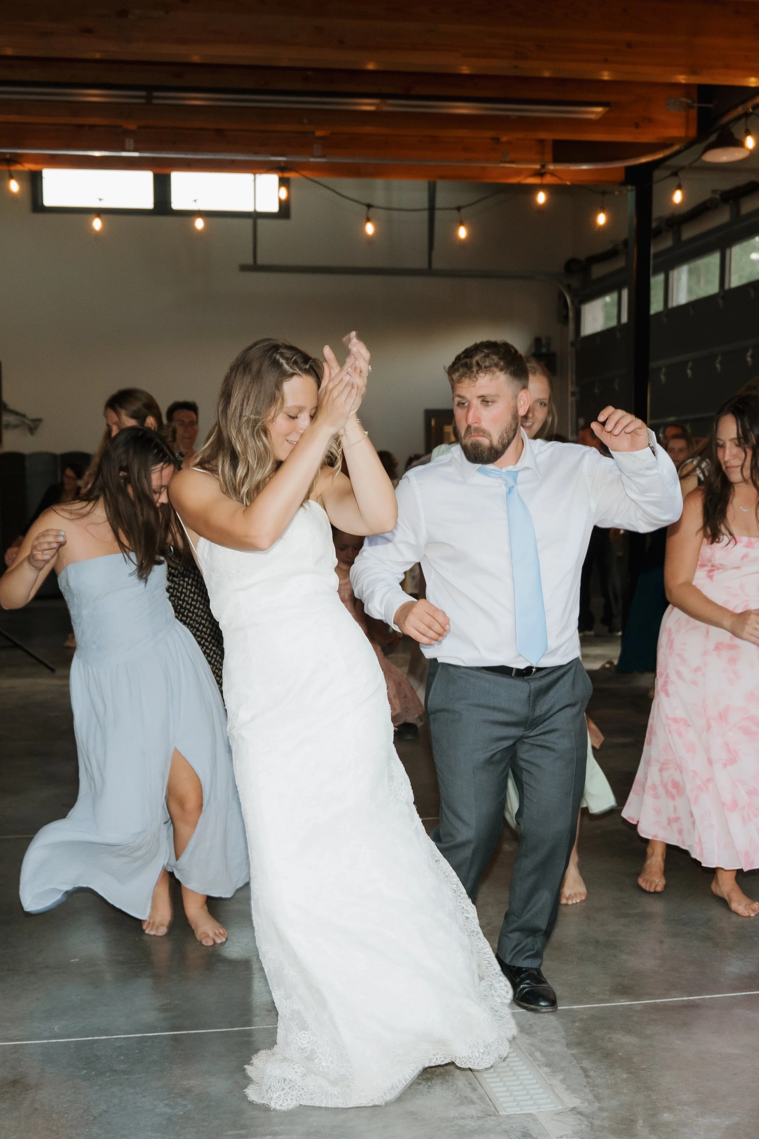 People dancing at a wedding reception, including a bride in a white wedding dress and a groom in a white shirt and blue tie, in a modern indoor venue.