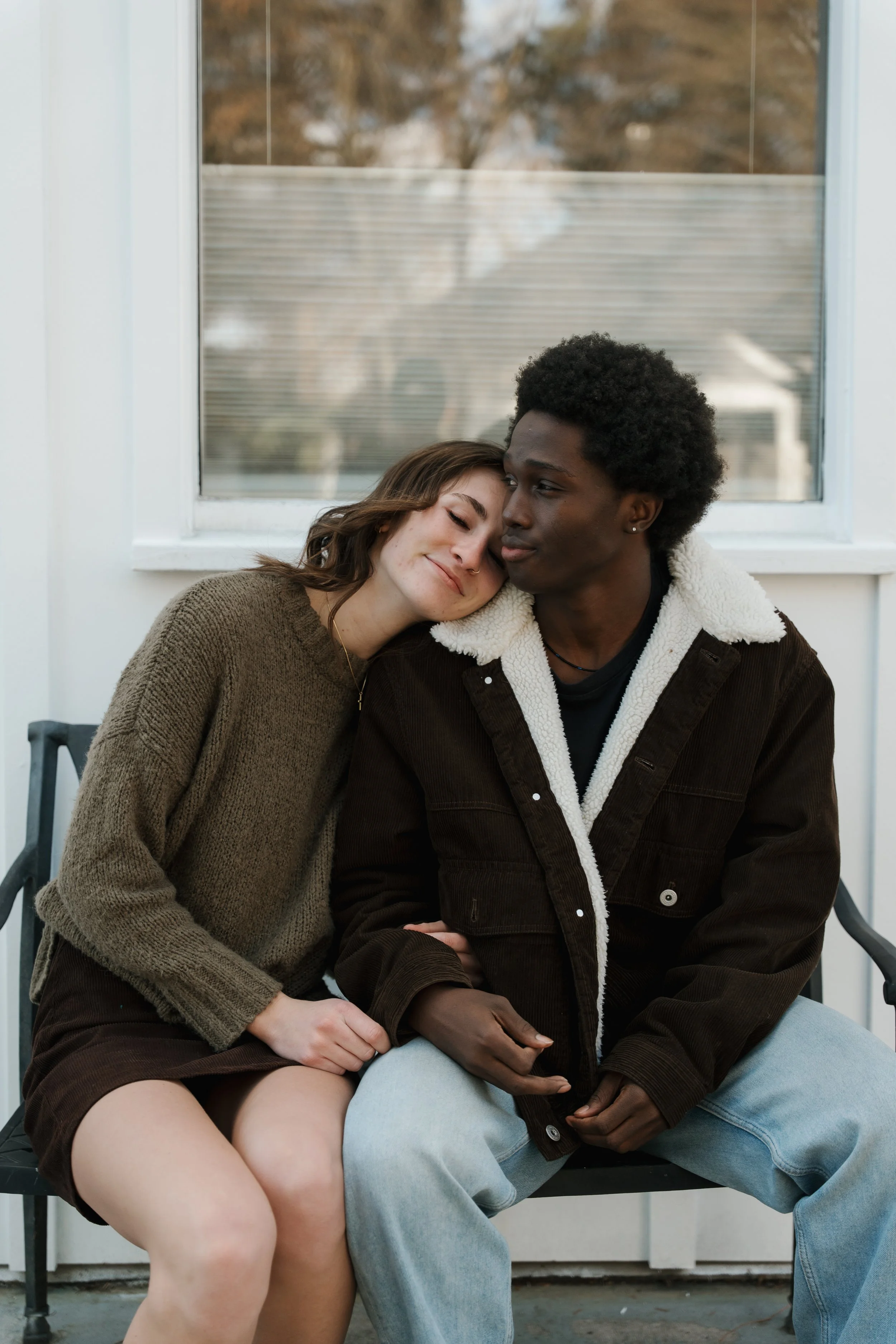 A young woman with brown hair leaning her head on a young man with black curly hair, sitting cozily together on a bench outside near a window.