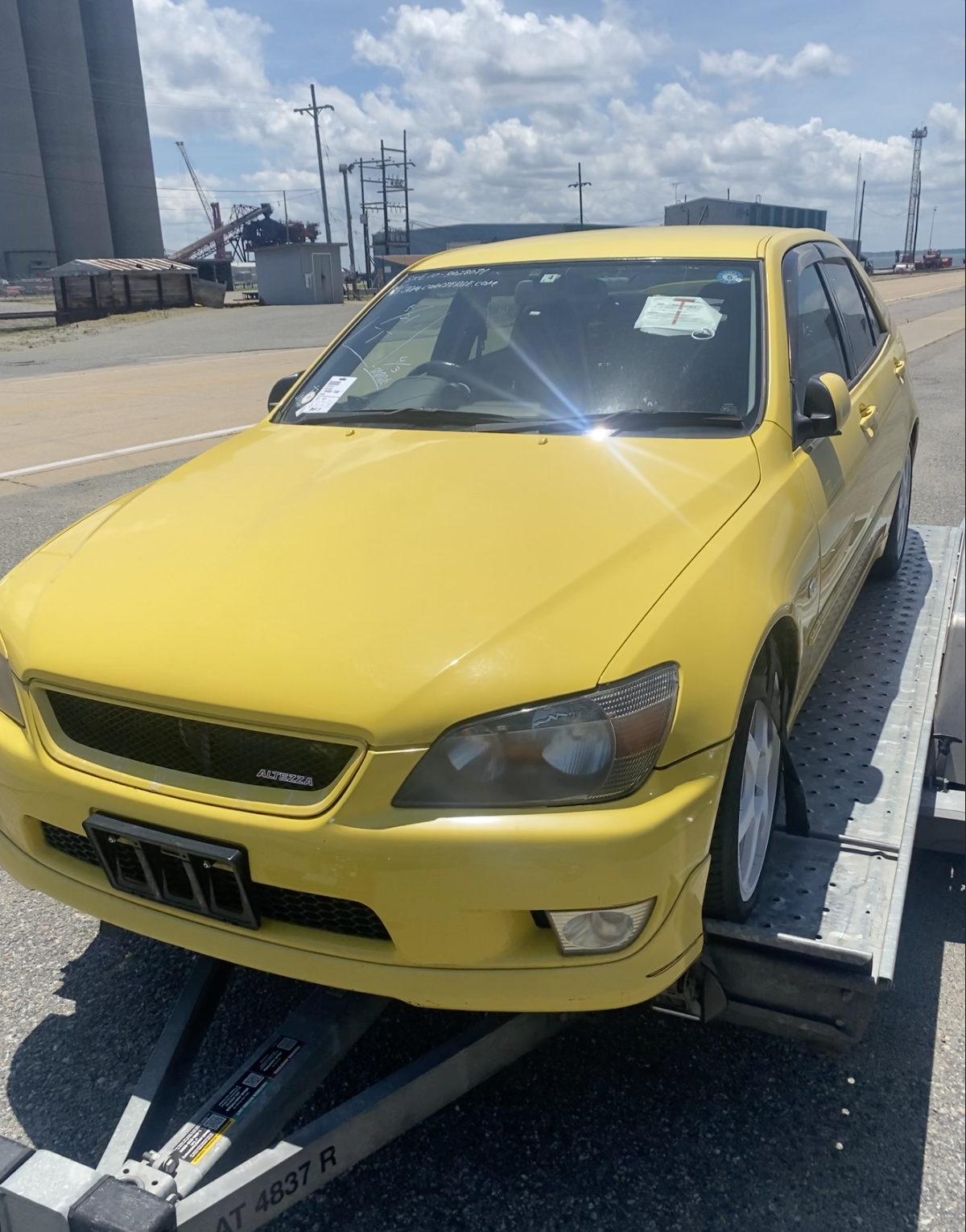 Yellow car on a trailer in an industrial area with blue sky and clouds.