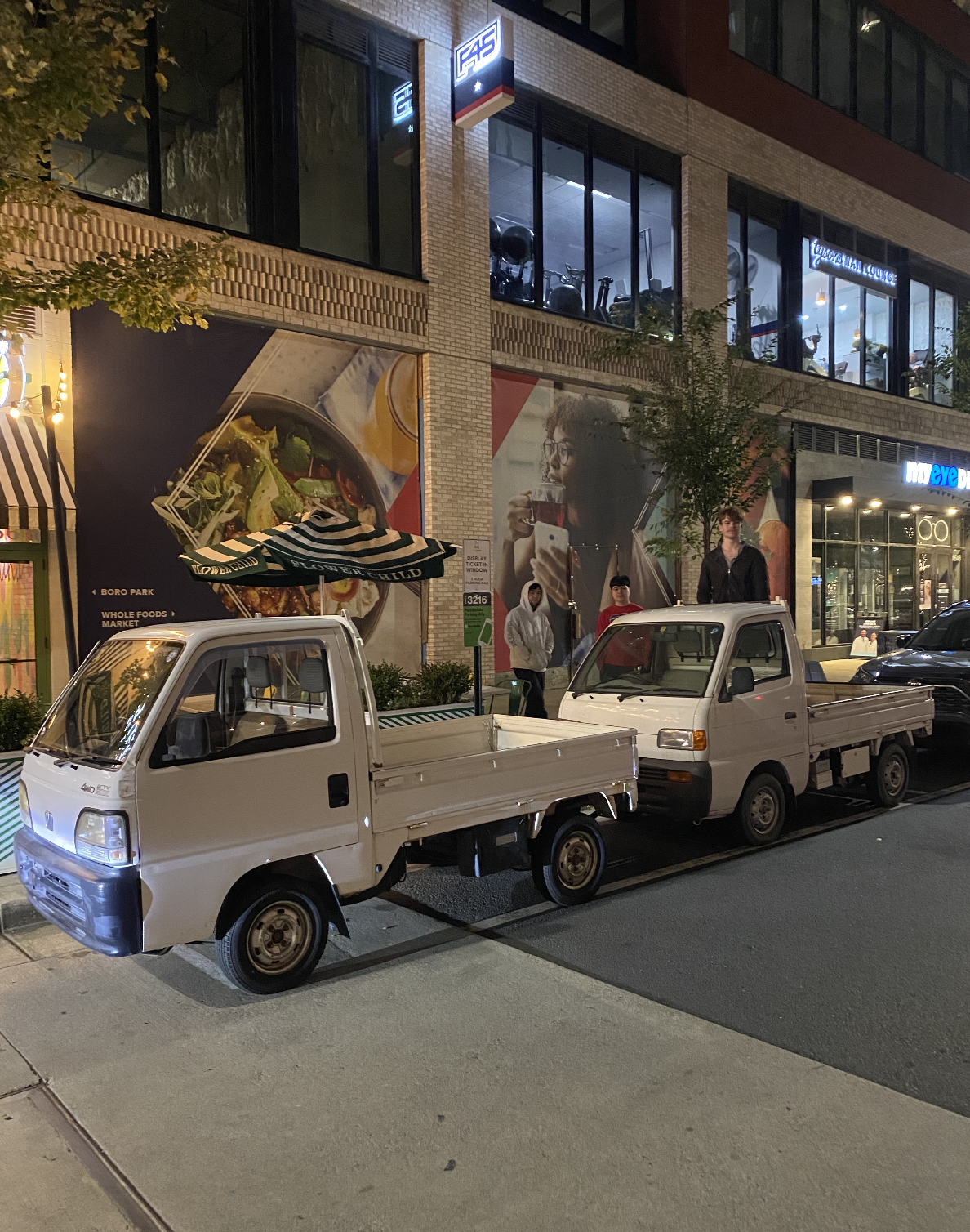 Two small white pickup trucks parked on a city street at night with storefronts in the background.