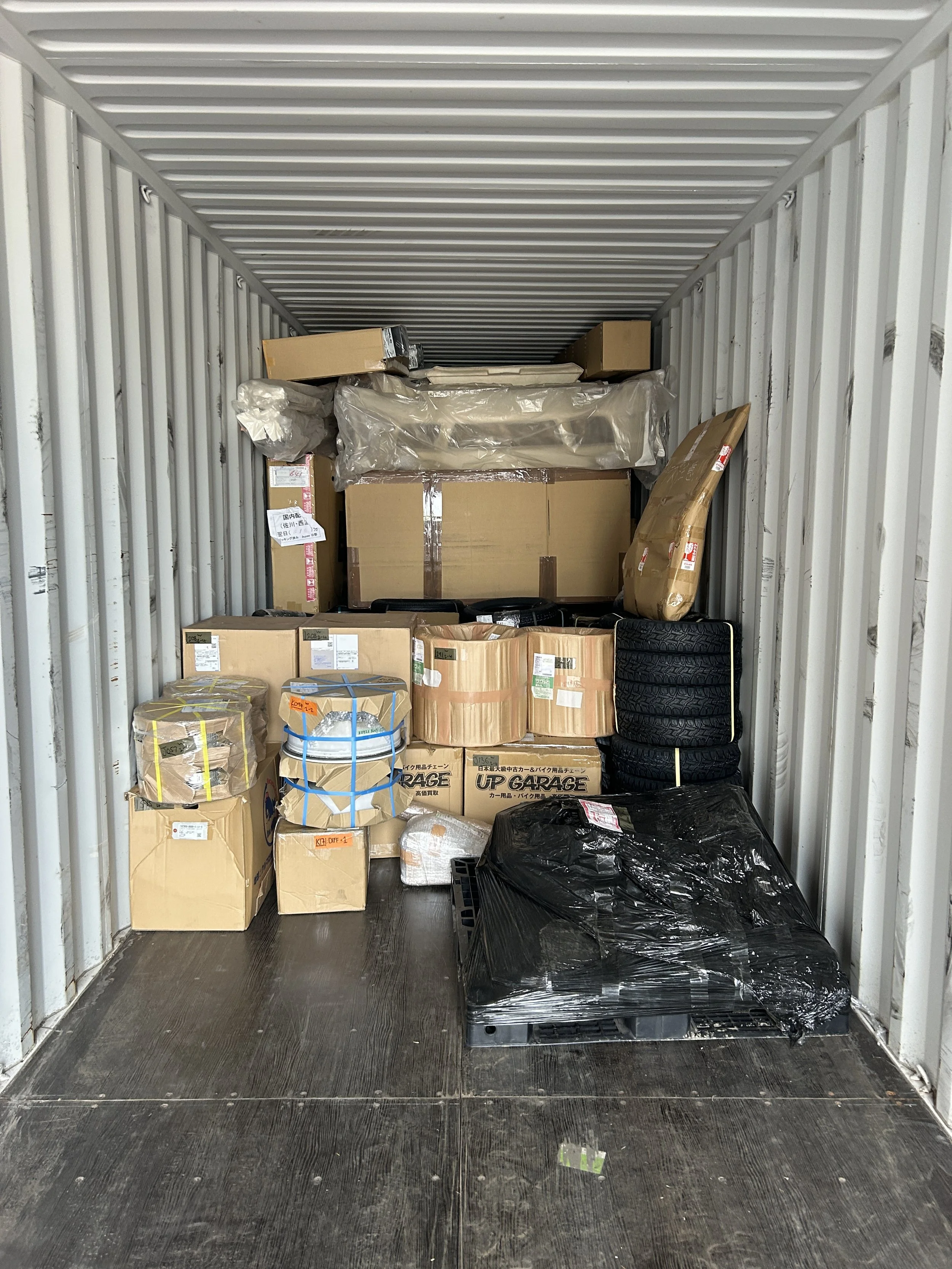 Inside a shipping container filled with stacked cardboard boxes, wrapped packages, and tires, with a dark wooden floor and metal walls.