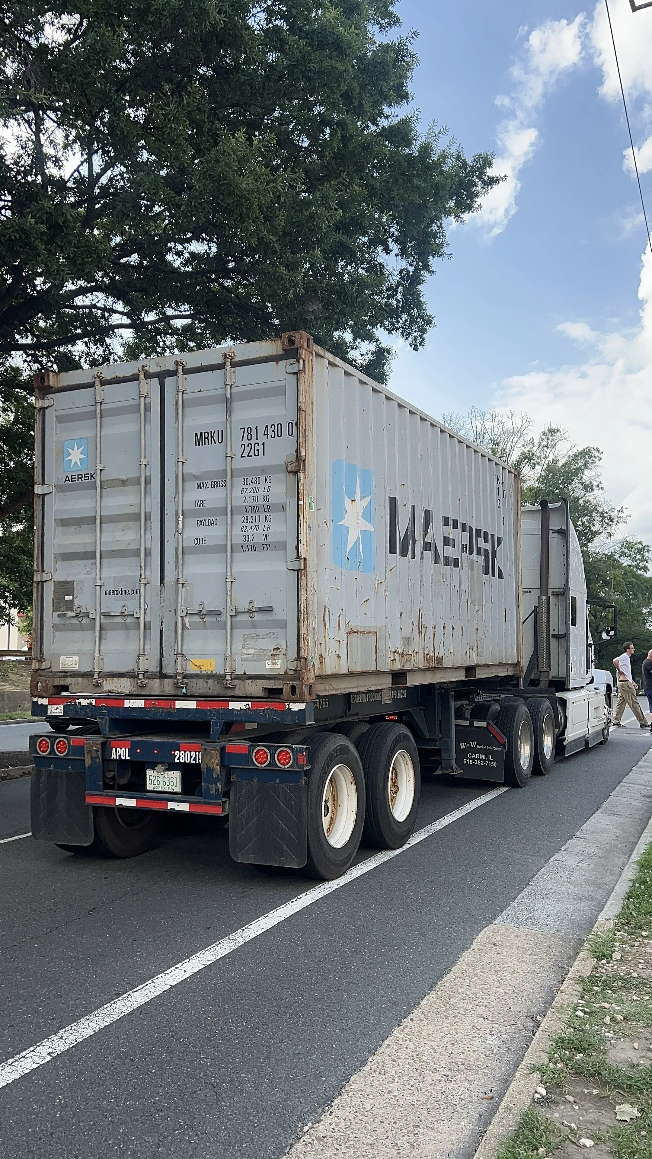 A gray semi-truck with a shipping container attached, parked on a street with a tree in the background and a person walking nearby.