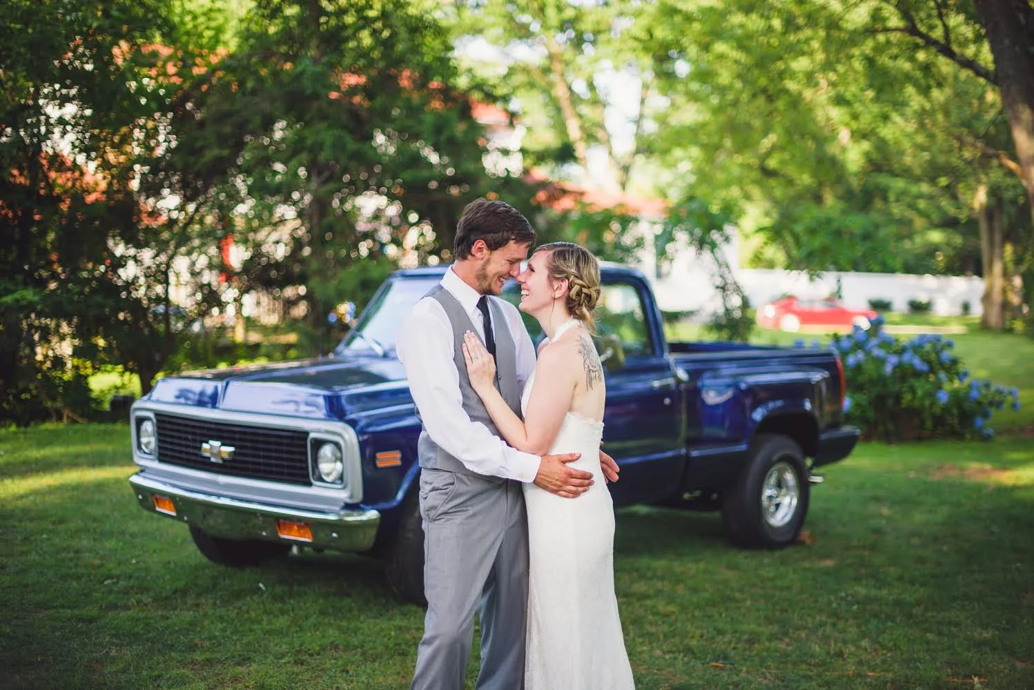 Bride and groom embracing in front of a classic blue pickup truck in a lush garden setting.