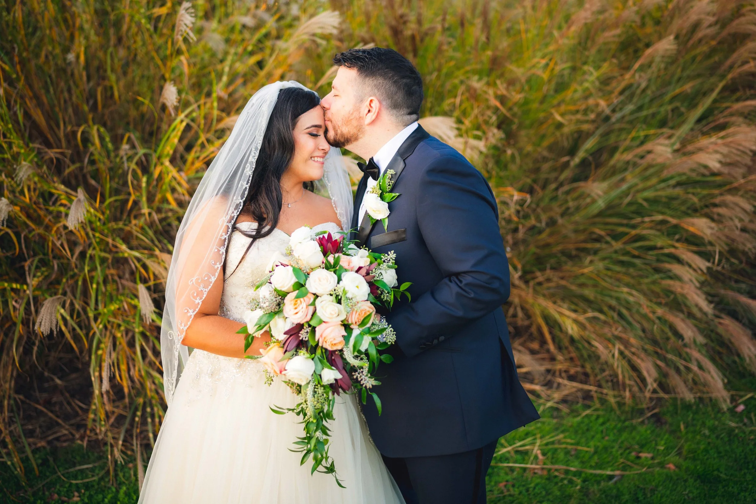 Bride and groom embracing outdoors, bride holding floral bouquet, grass in background.