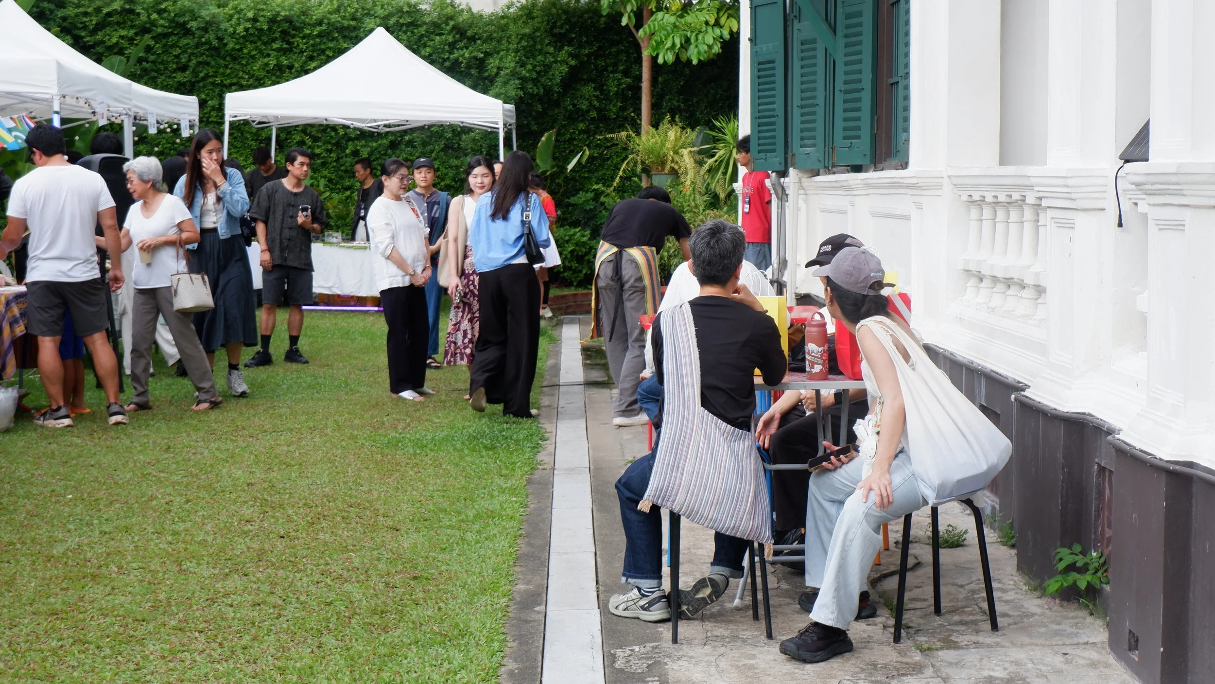 People gathered outdoors at a market or event, standing and sitting near a white building with green shutters, green trees in the background, and white tents overhead.