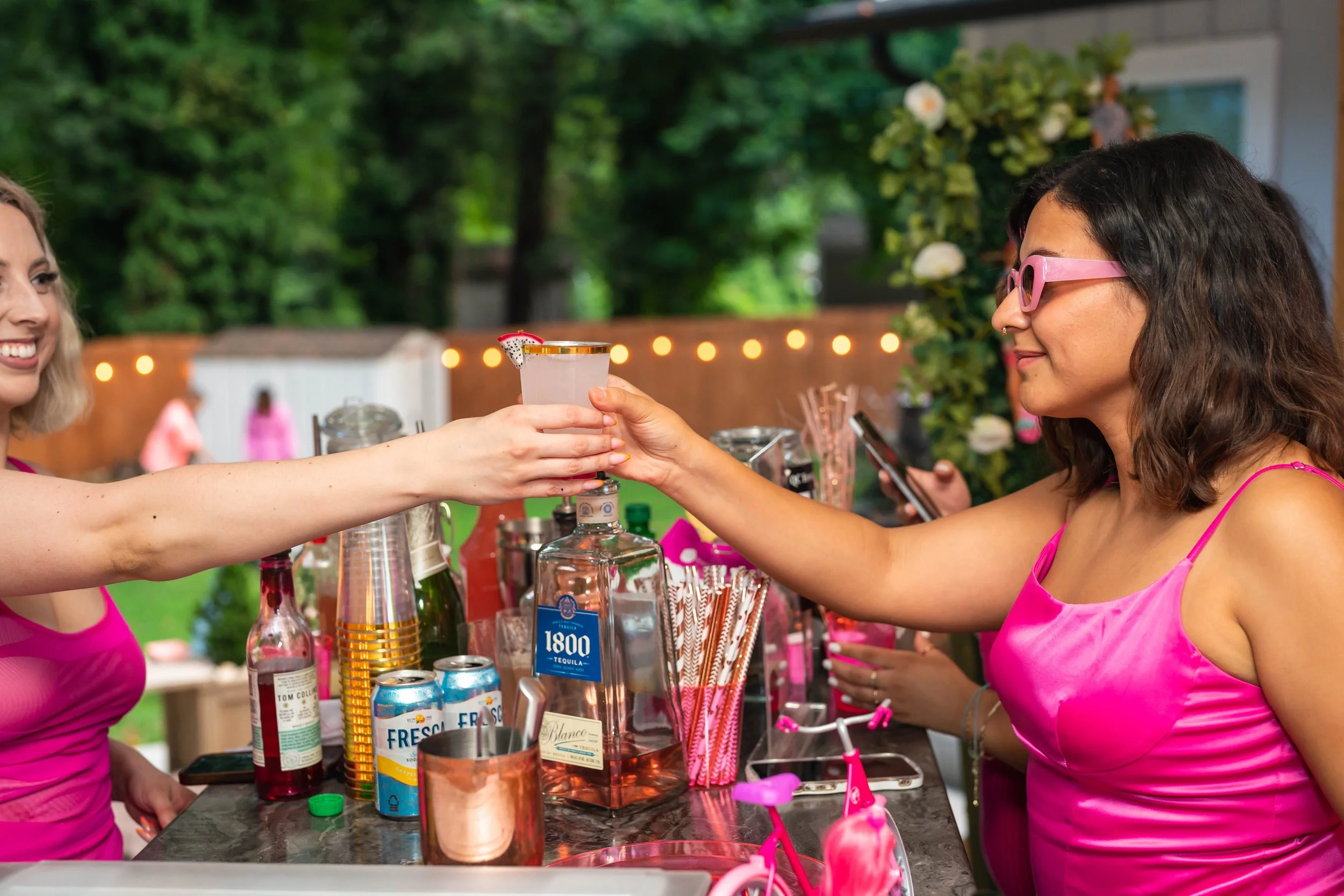 Two women in pink tops enjoying drinks at an outdoor bar with bottles and cans on the counter.