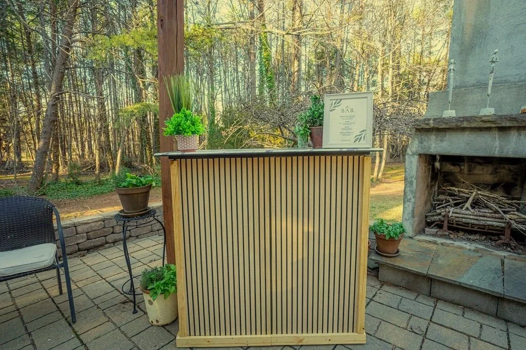 Outdoor bar setup on a patio with potted plants, a chair, and a stone fireplace under a wooden pergola, surrounded by trees.
