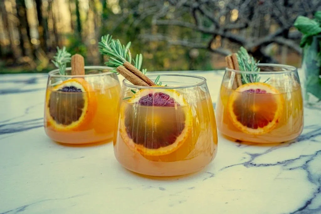 Three glasses of orange-colored cocktails garnished with orange slices, cinnamon sticks, and rosemary sprigs on a marble surface with a blurred forest background.