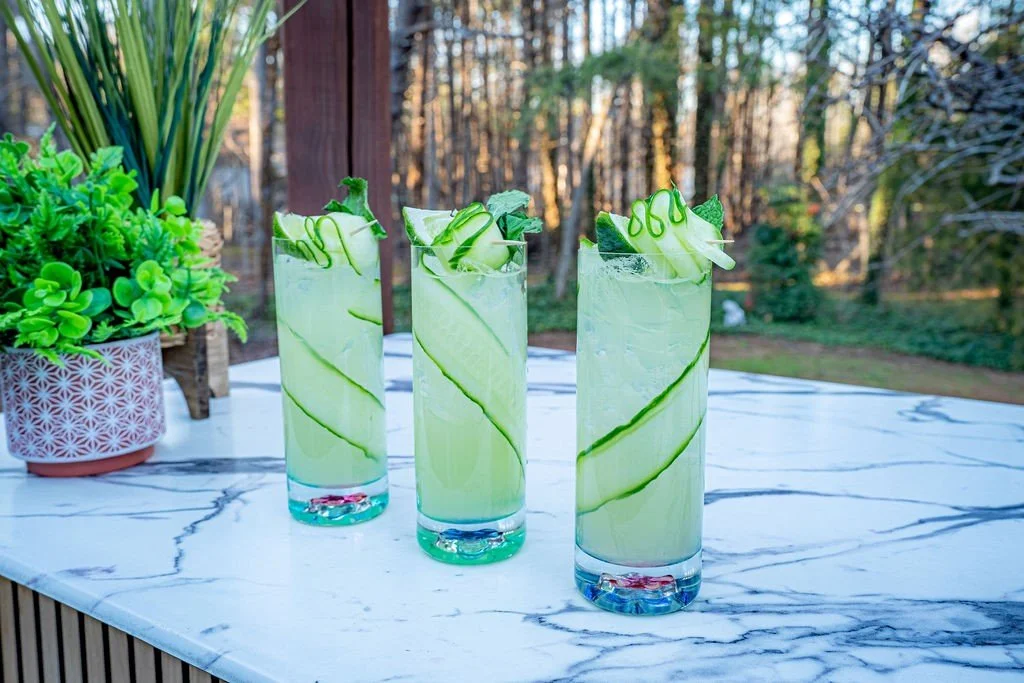 Three glasses of cucumber-infused drinks on a marble table near a potted plant.