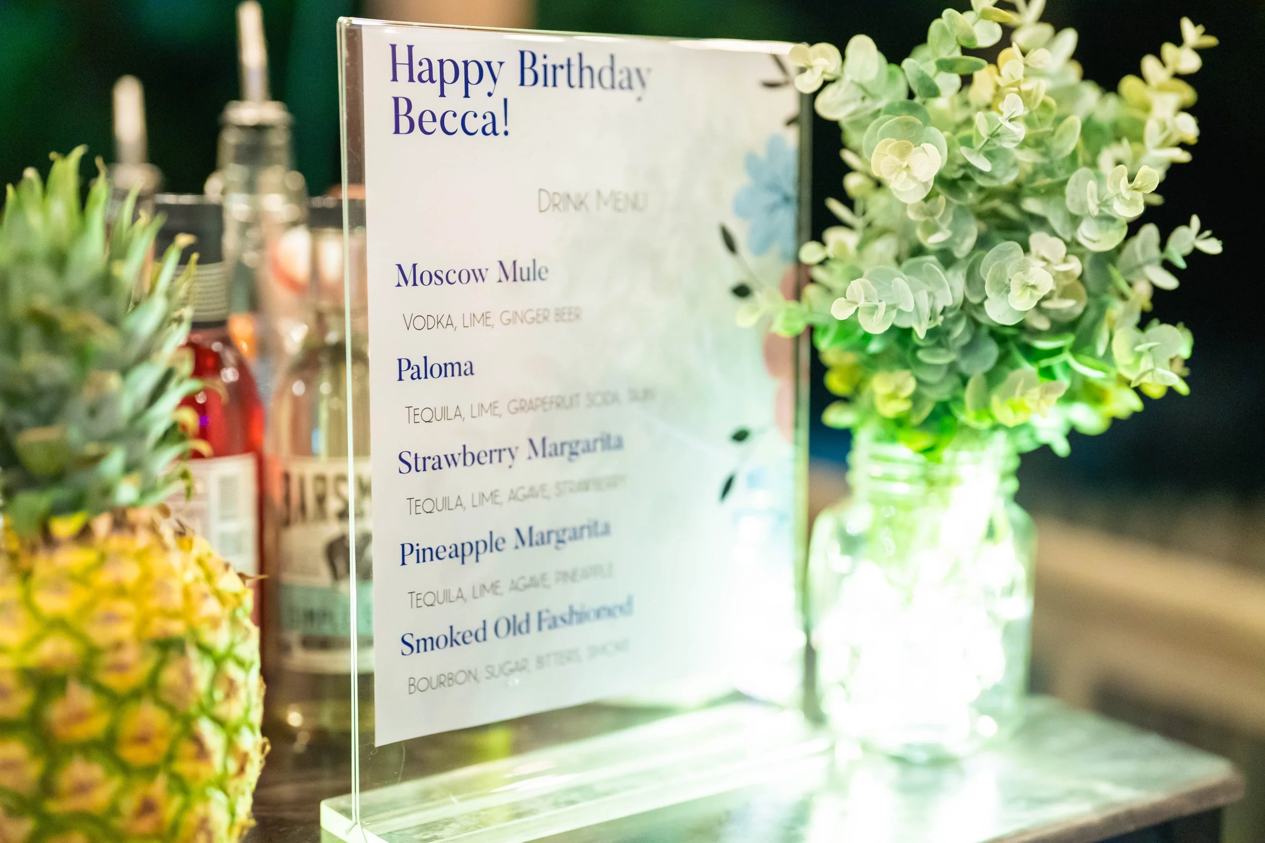 Birthday party drink menu with various cocktails, featuring a decorative arrangement of green foliage and glass bottles in the background, alongside a pineapple.