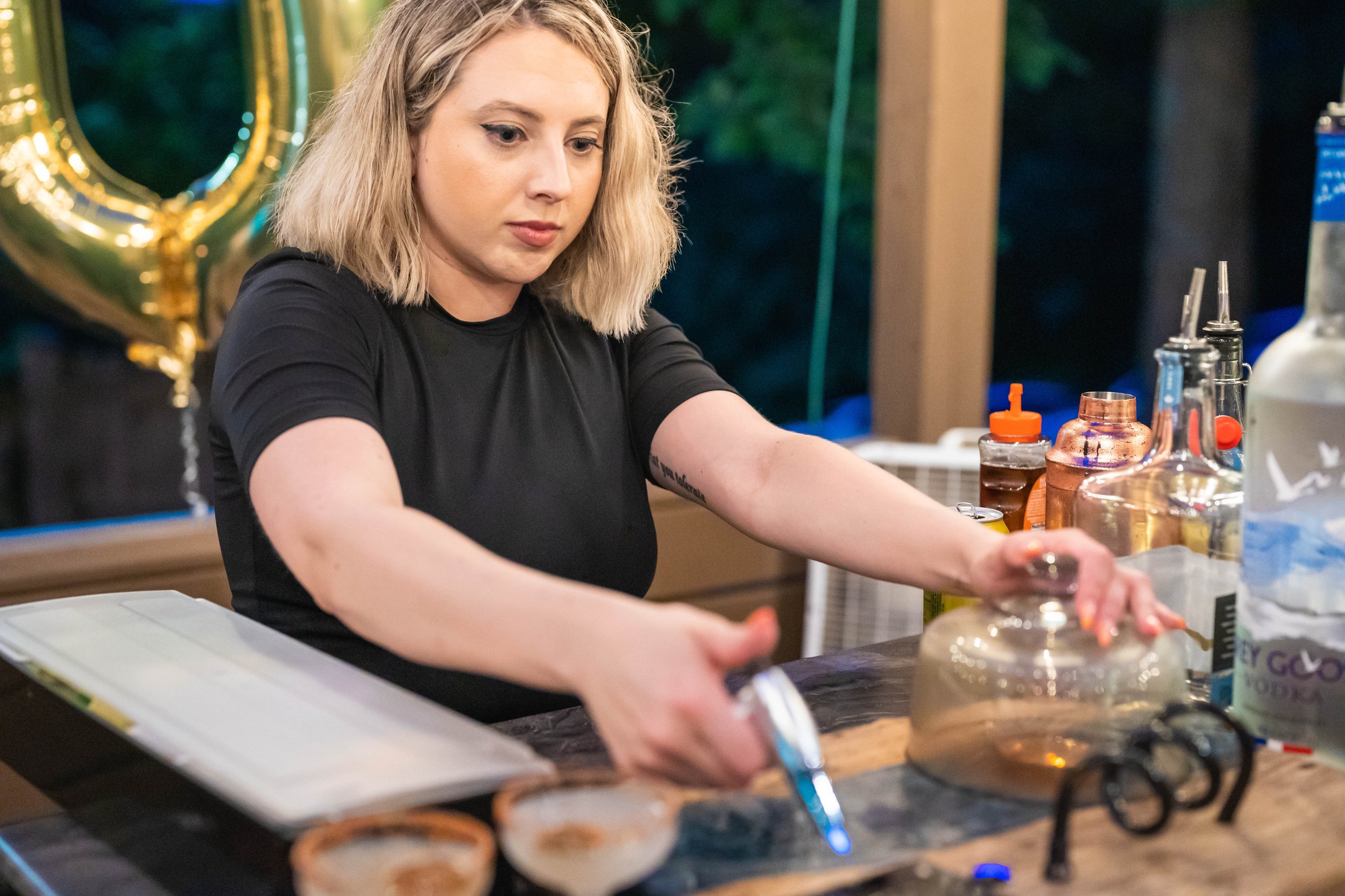 Person preparing cocktails at a bar with various bottles and a cocktail shaker.