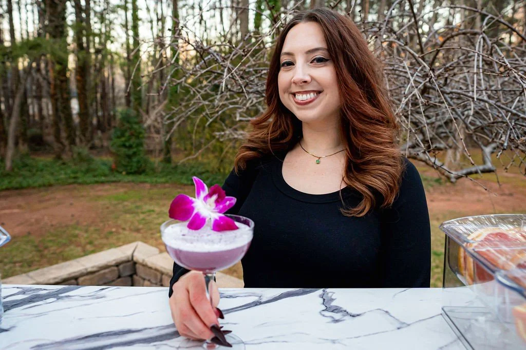 Smiling woman holding a cocktail with a purple flower garnish at an outdoor setting.