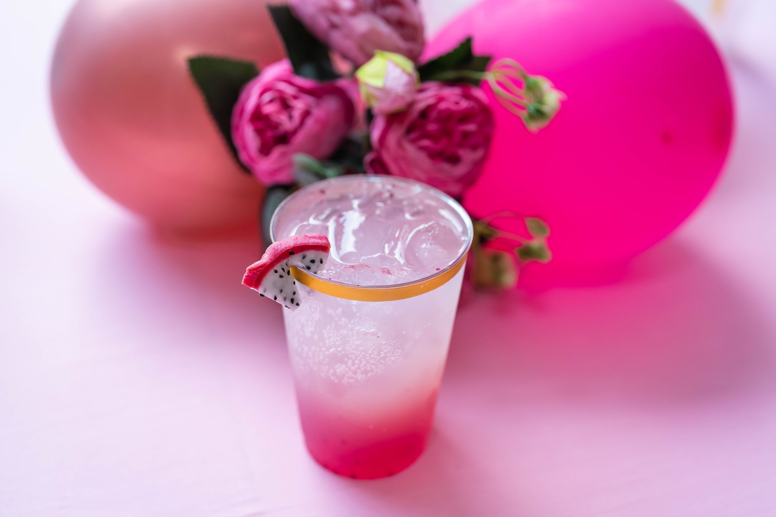 Pink drink with ice in a glass, garnished with dragon fruit slice, surrounded by pink balloons and flowers.