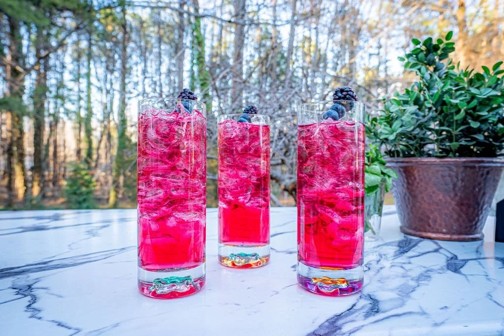 Three tall glasses filled with a bright pink beverage garnished with berries, placed on a marble surface with outdoor greenery in the background.