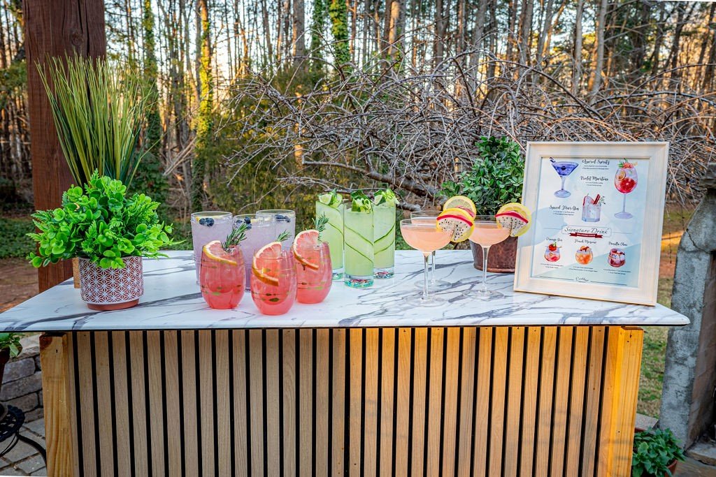 Outdoor bar with various cocktails, framed drink menu, and potted plants on a marble countertop.