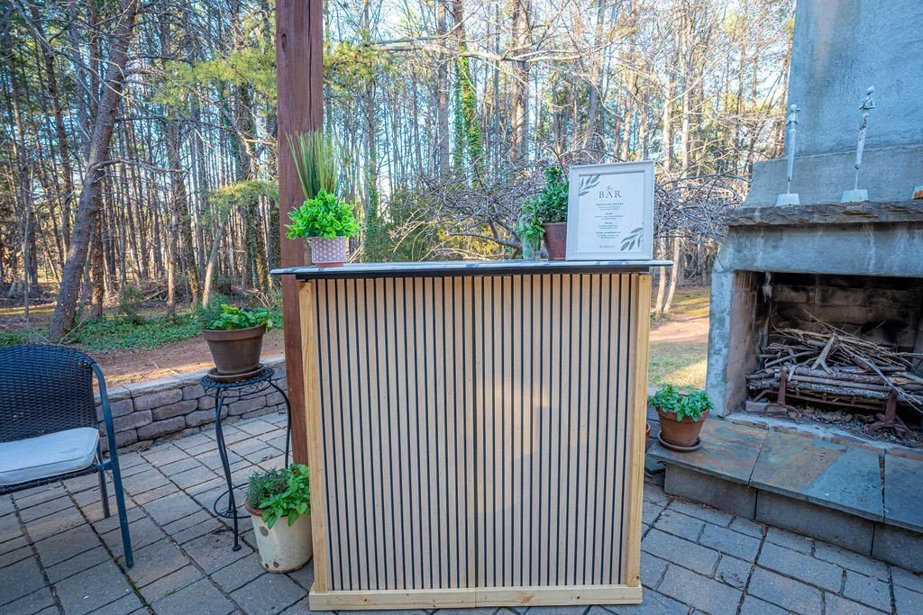 Outdoor bar setup on a patio with potted plants, a chair, and a stone fireplace under a wooden pergola, surrounded by trees.