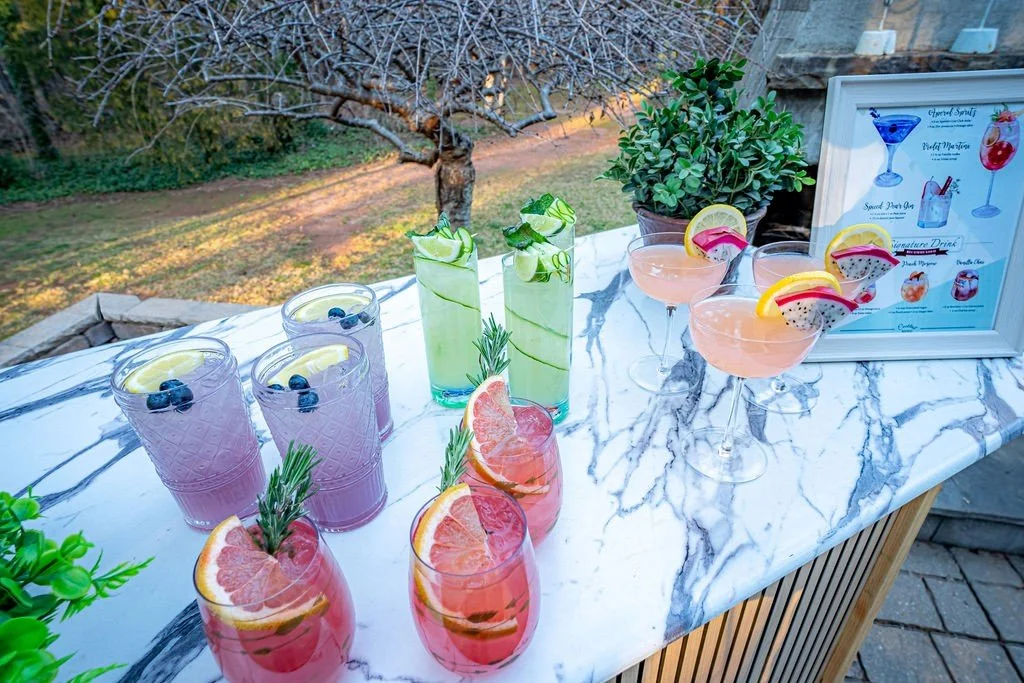 Colorful cocktails on a marble table outdoors, featuring drinks garnished with fruit slices and herbs, with a menu and potted plants in the background.