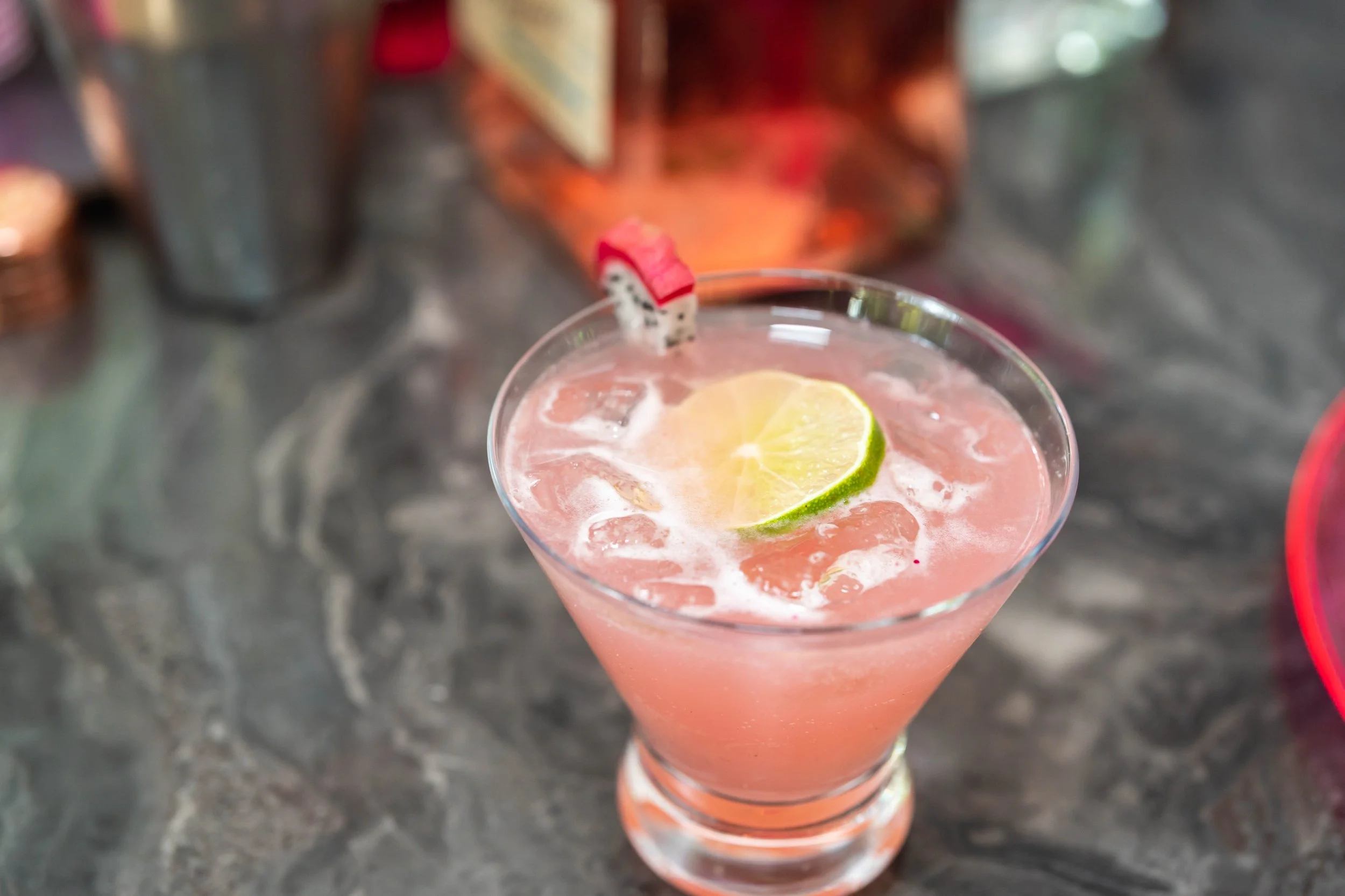 Pink cocktail with ice, lime slice, and dragon fruit garnish in a glass on a dark marble surface.