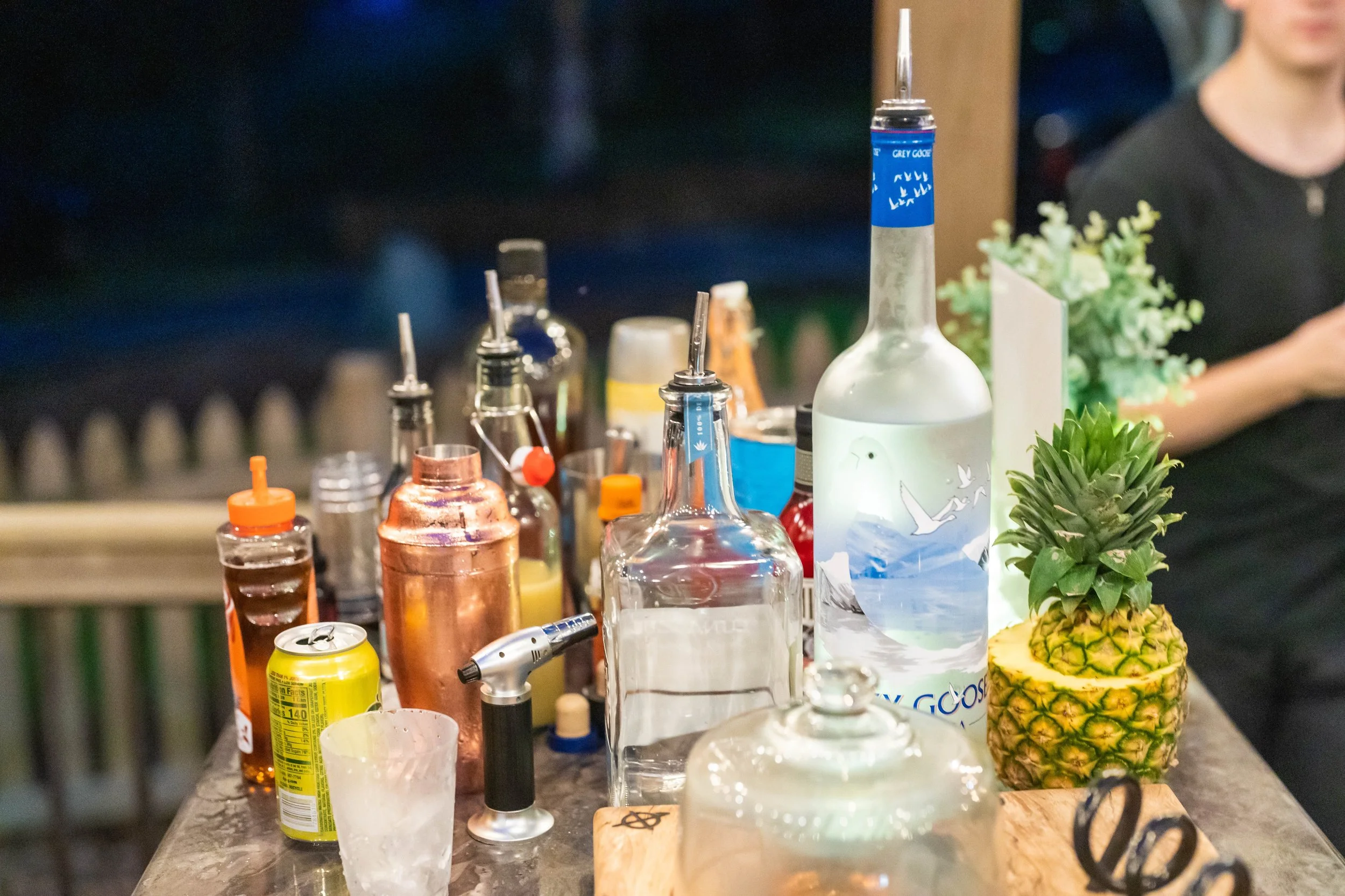Bar setup with alcohol bottles, a cocktail shaker, a sliced pineapple, and bartending tools on a counter.