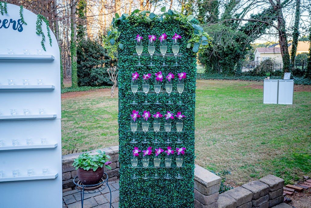 Outdoor champagne wall display with green foliage, holding glasses of champagne with purple flowers, on a patio with brick border, and trees in the background.