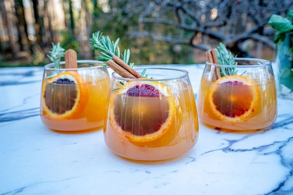Three glasses of orange-colored cocktails garnished with orange slices, cinnamon sticks, and rosemary sprigs on a marble surface with a blurred forest background.