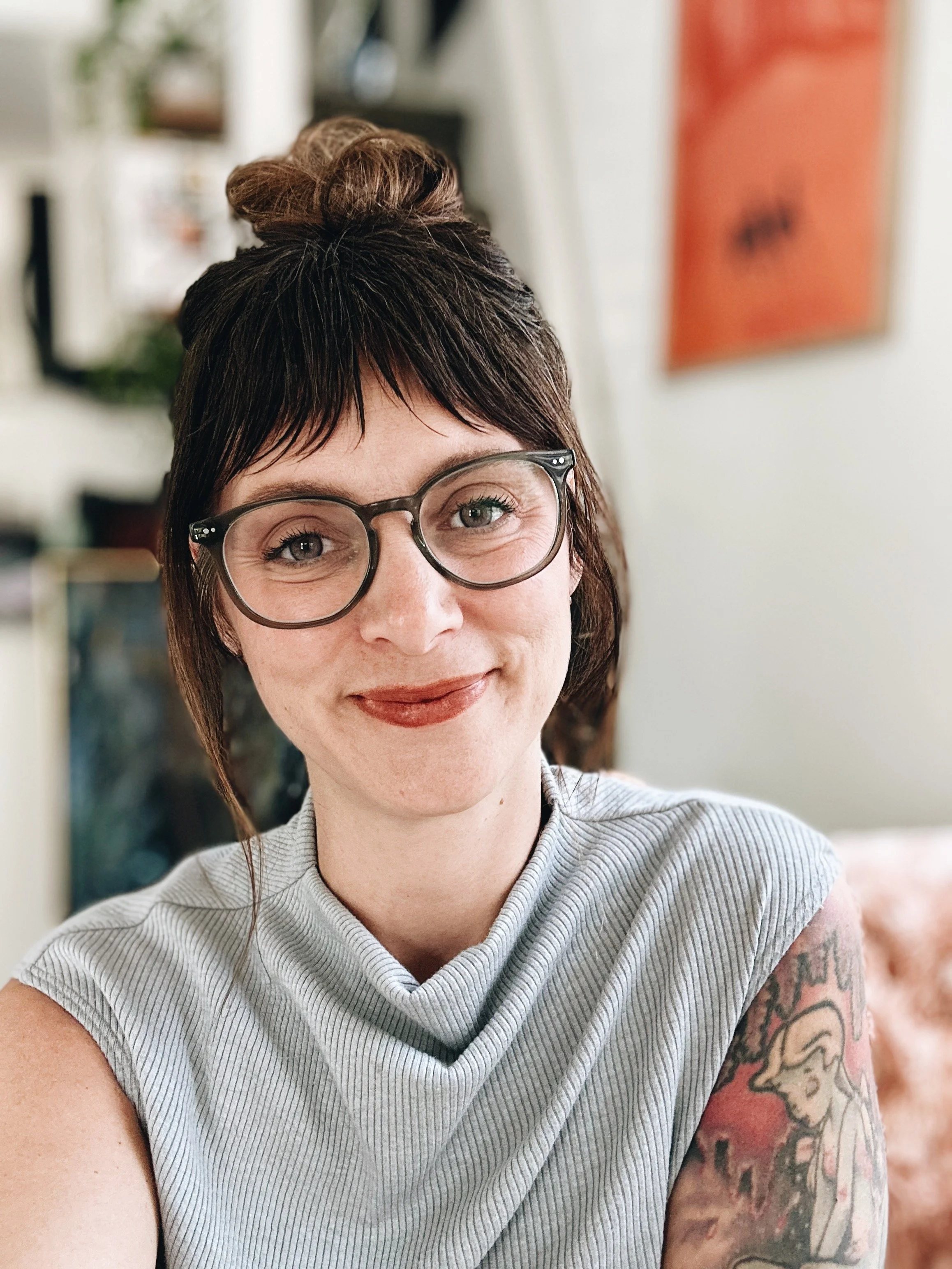 A woman with short dark hair styled in a messy bun, wearing large round glasses, smiling at the camera. She is wearing a light gray, striped, short-sleeve top and has a colorful tattoo sleeve on her right arm. A portrait of a mental health therapist.