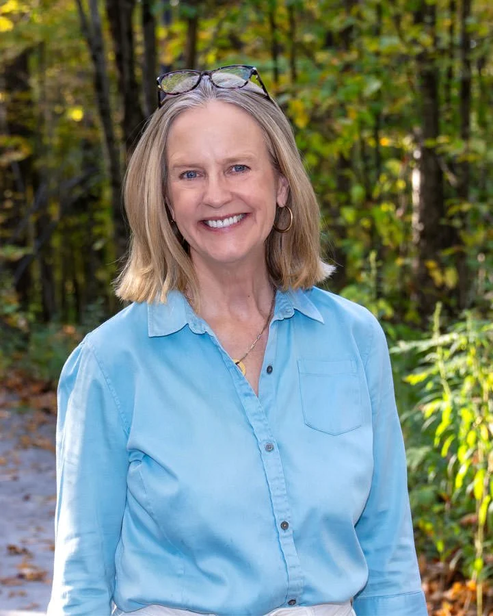 Portrait of a woman standing amongst fall foliage and smiling. She has her glasses on her head. 
