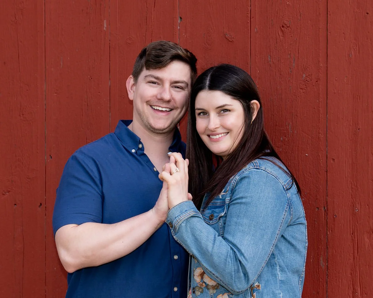couple standing close together in front of a red barn wall having just got engaged