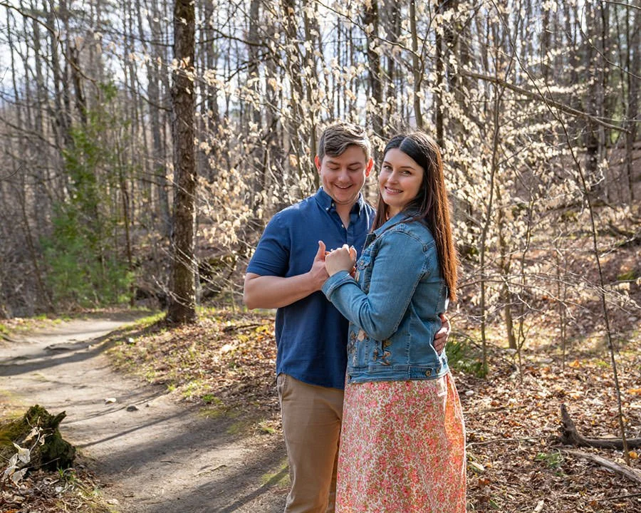a couple who are just engaged stand together in front of trees lit by low sunlight