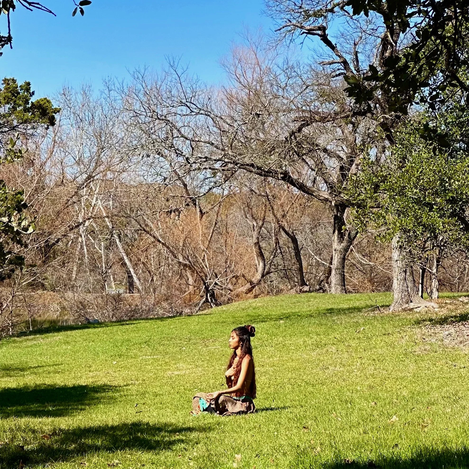A person meditating on a green grassy field surrounded by leafless trees and some with green foliage under a clear blue sky.