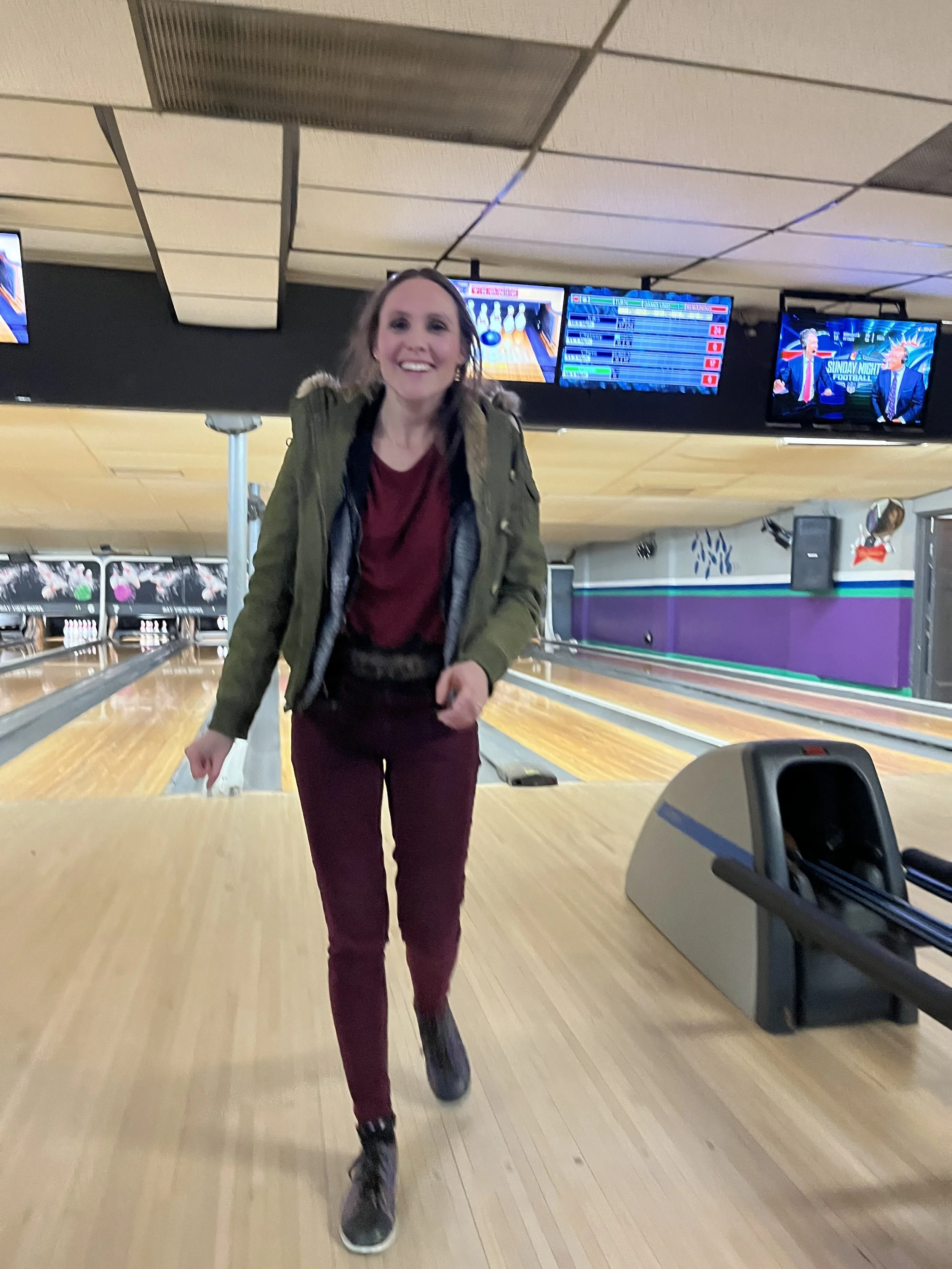 A woman in a green jacket and maroon pants is bowling, approaching the camera with a smile, in a bowling alley with several screens and bowling lanes.