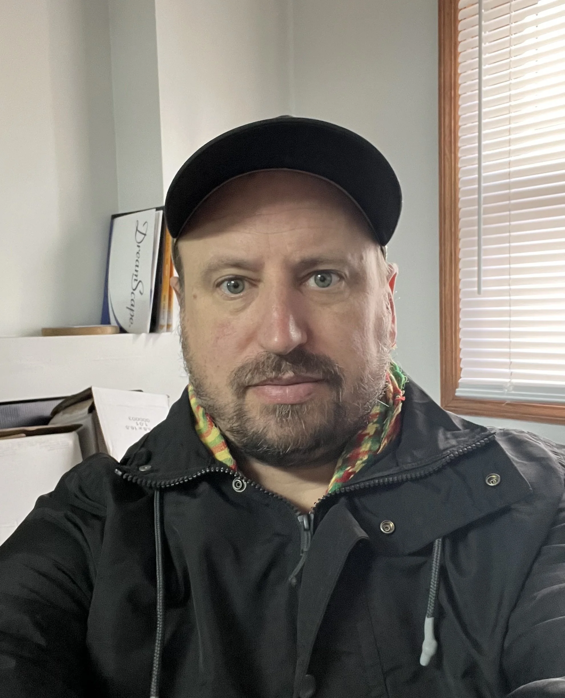 A man with a beard and light skin wearing a black baseball cap, black jacket, and a colorful hoodie taking a selfie indoors. Behind him is a window with white blinds, a white wall, and a shelf with books and a framed certificate.