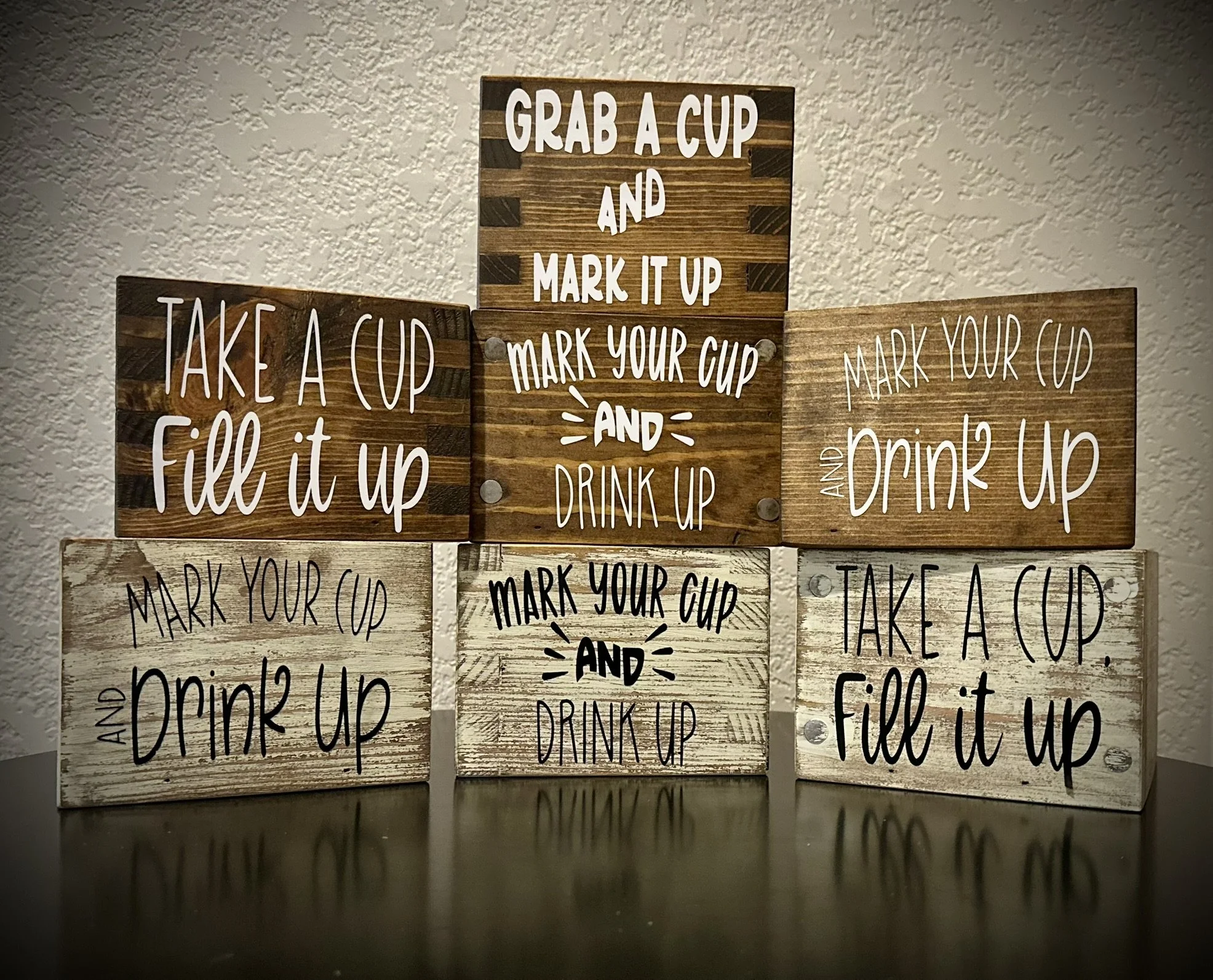 Six wooden signs with drink-related instructions and decorative fonts, displayed on a dark surface against a textured wall. The signs have phrases like "Grab a cup and mark it up," "Take a cup fill it up," and "Mark your cup and drink up."