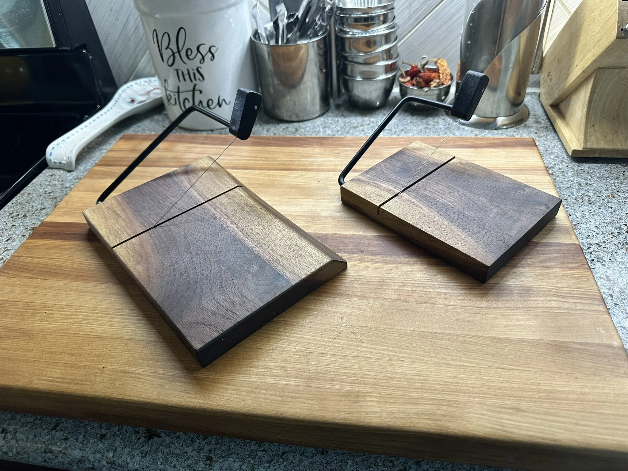 Two wooden cutting boards with black handles and metal rods are placed on a kitchen counter. The counter has various kitchen items, including jars, a pot, and a paper towel holder with the words 'Bless this Kitchen' written on it.