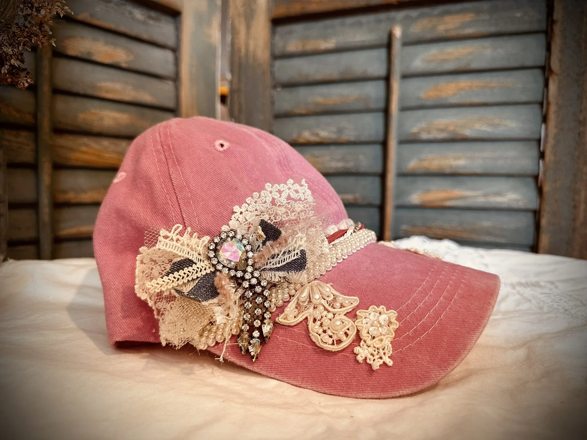 A pink cap decorated with lace, pearls, embroidery, and a large jewel embellishment on the front, placed on a white surface with a wooden fence background.
