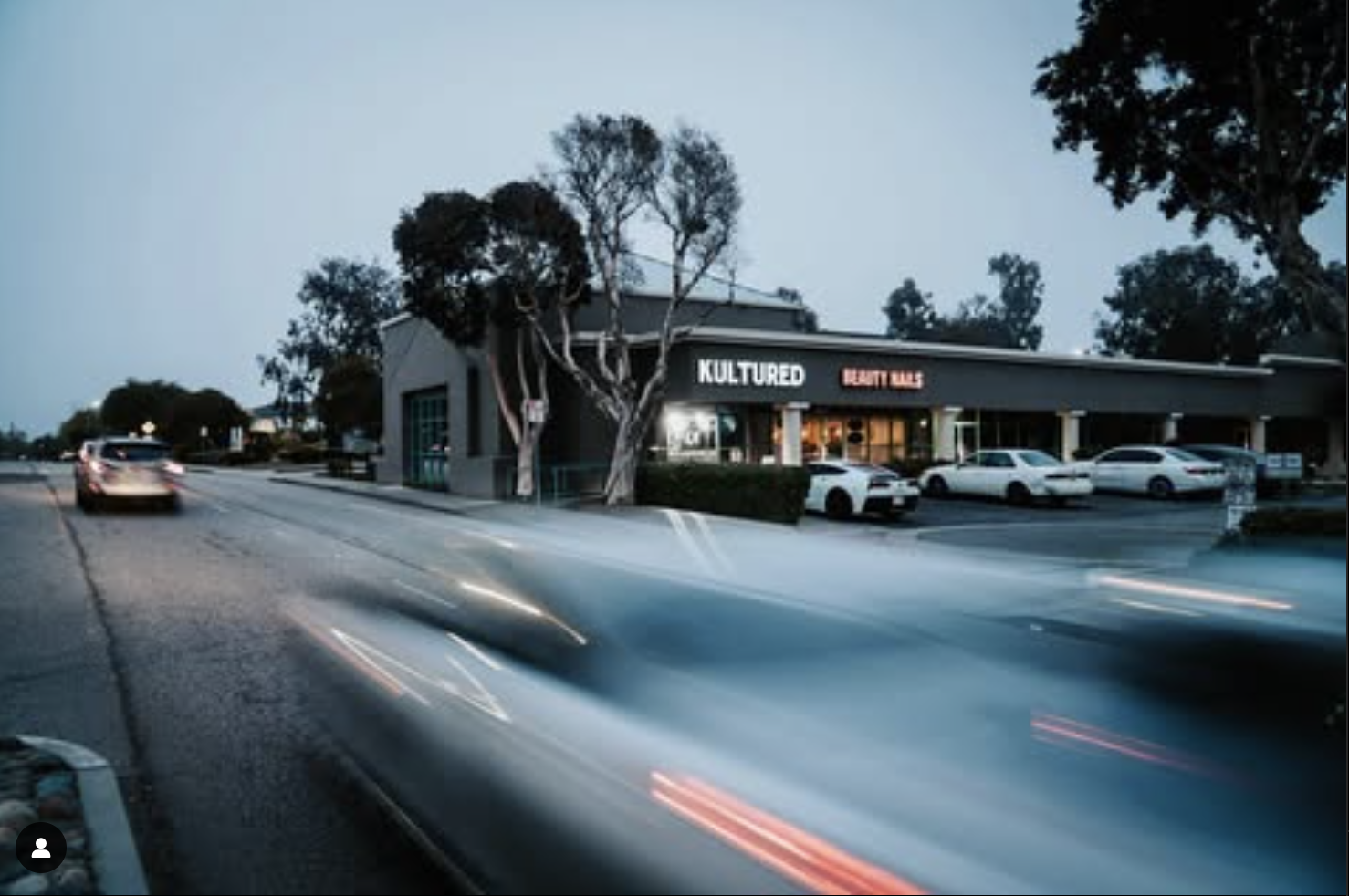 Street scene with cars passing by a building in twilight, featuring "Kultured Beauty Nails" signage. Trees and parked cars are visible in the background.
