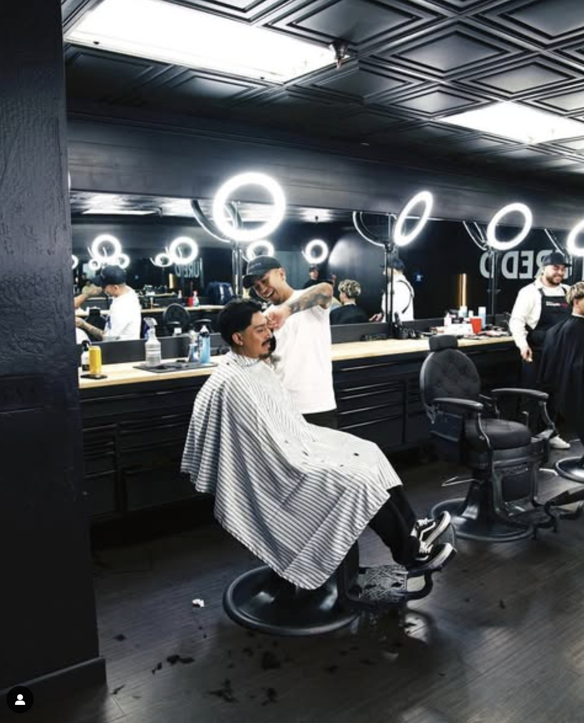 Barber shop interior with a barber cutting a customer's hair under bright ring lights, surrounded by mirrors and salon equipment.
