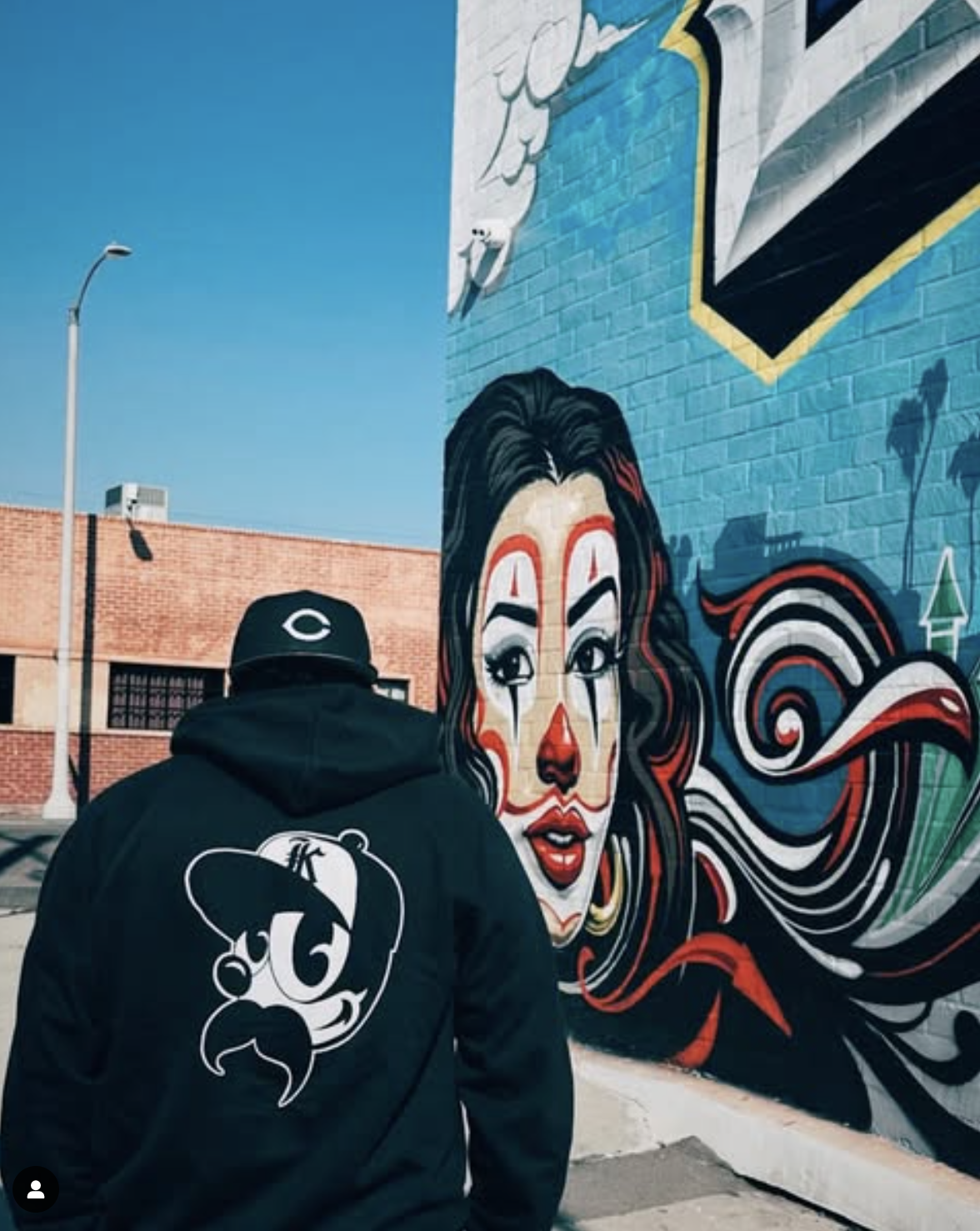Person in hoodie and cap standing in front of a colorful mural of a woman's face on a brick wall, depicting vibrant swirls and patterns, with urban street elements in the background.